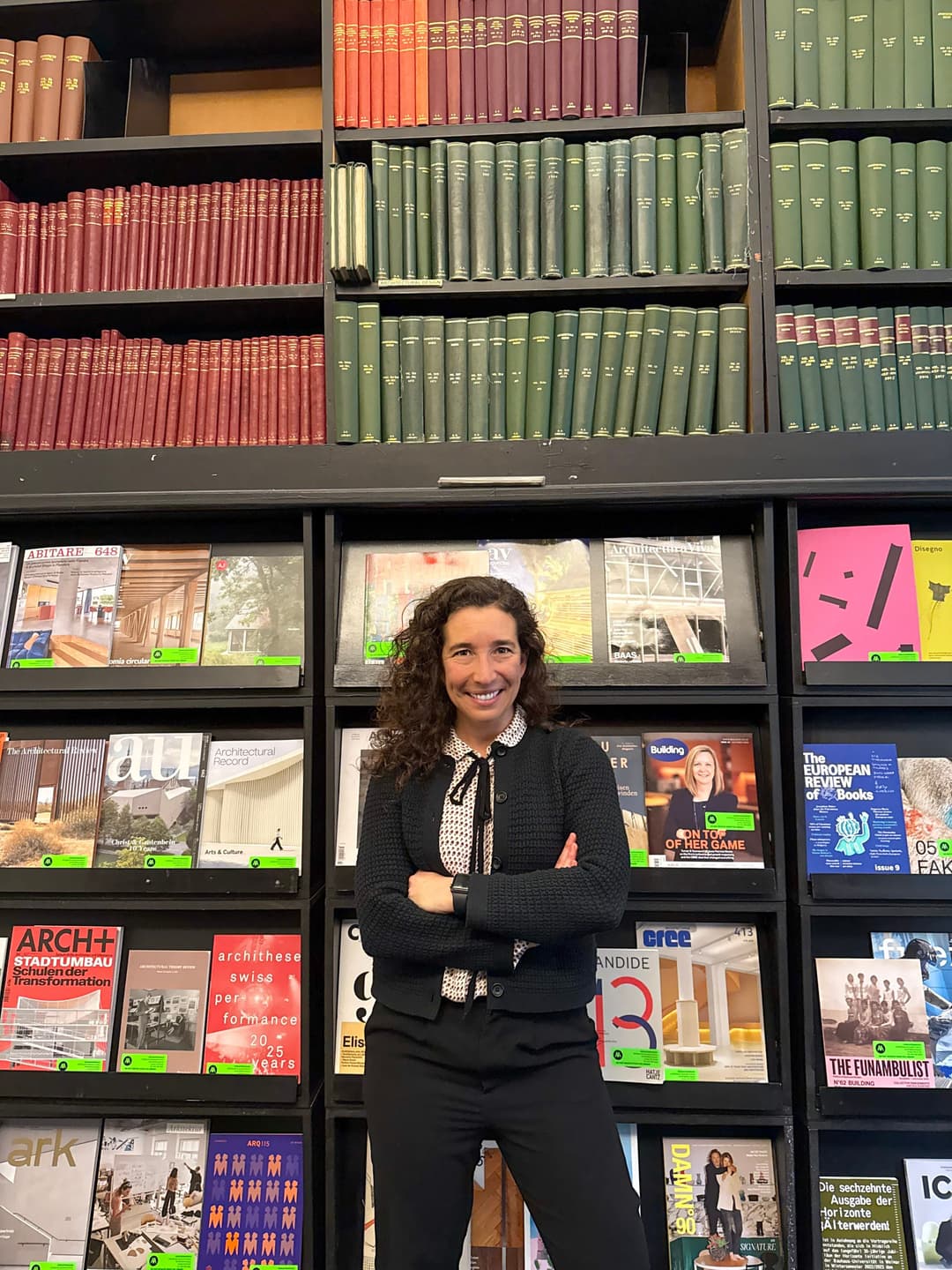 A woman with curly hair smiles, arms crossed, in front of a wall of books.