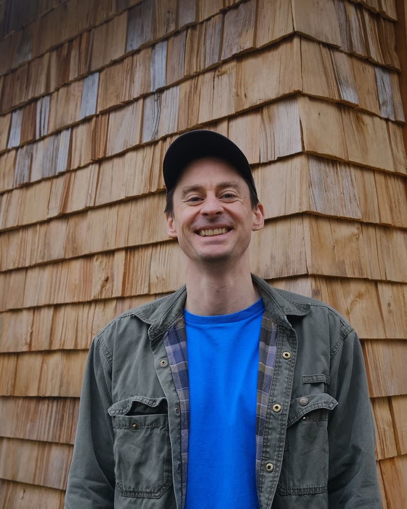 A white skinned man in a denim jacket, blue top and peaked cap stands in front of a timber shingled wall.