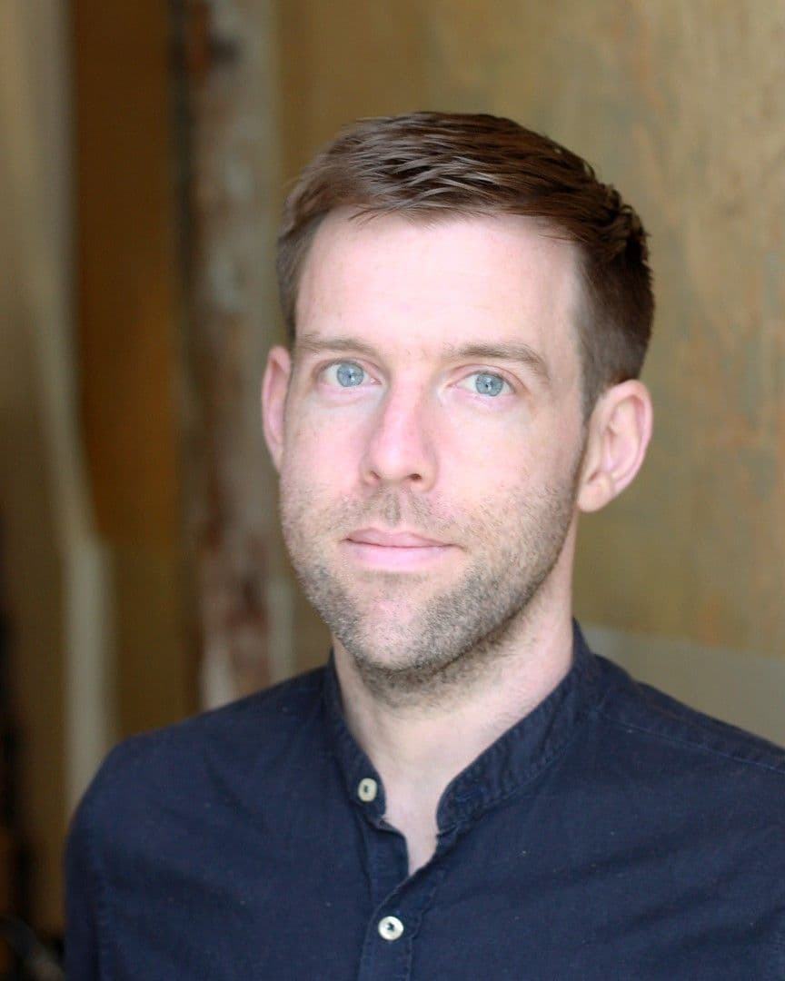 A man with short brown hair and a beard wears a black collared shirt.