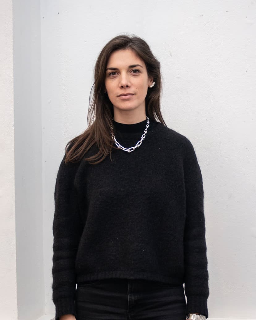 A young woman with long brown hair and dark clothing stands against a white wall.