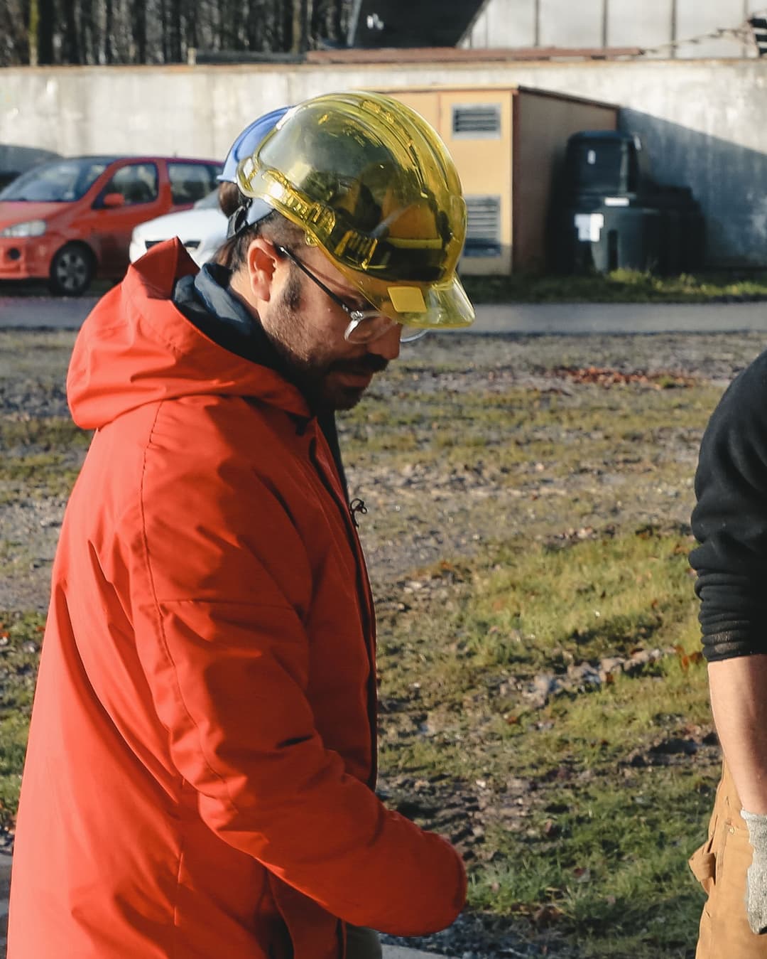A man wearing a yellow hard hat and a bright orange jacket stands in a working yard.