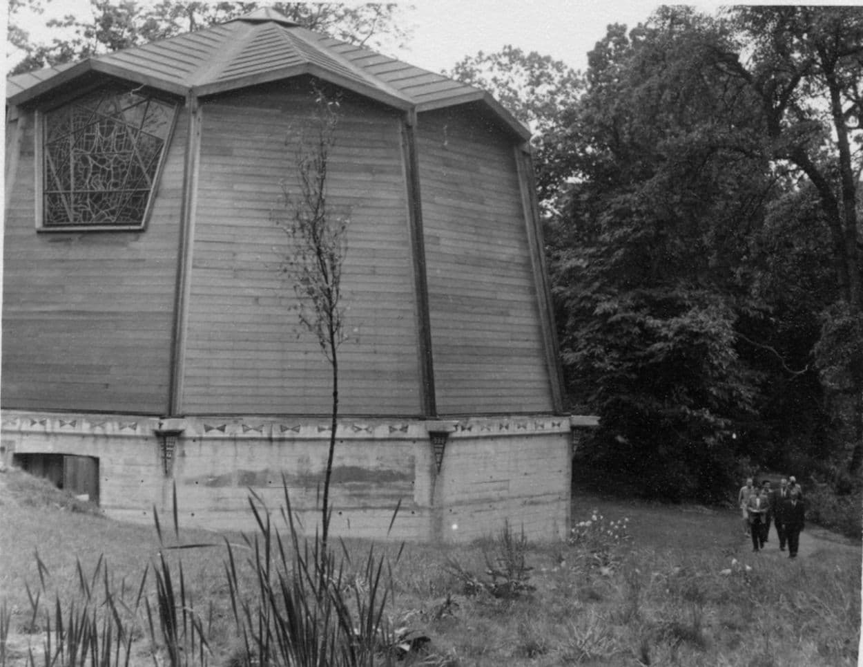 Octagonal wood building with geometric window nestled in a grassy, wooded area.