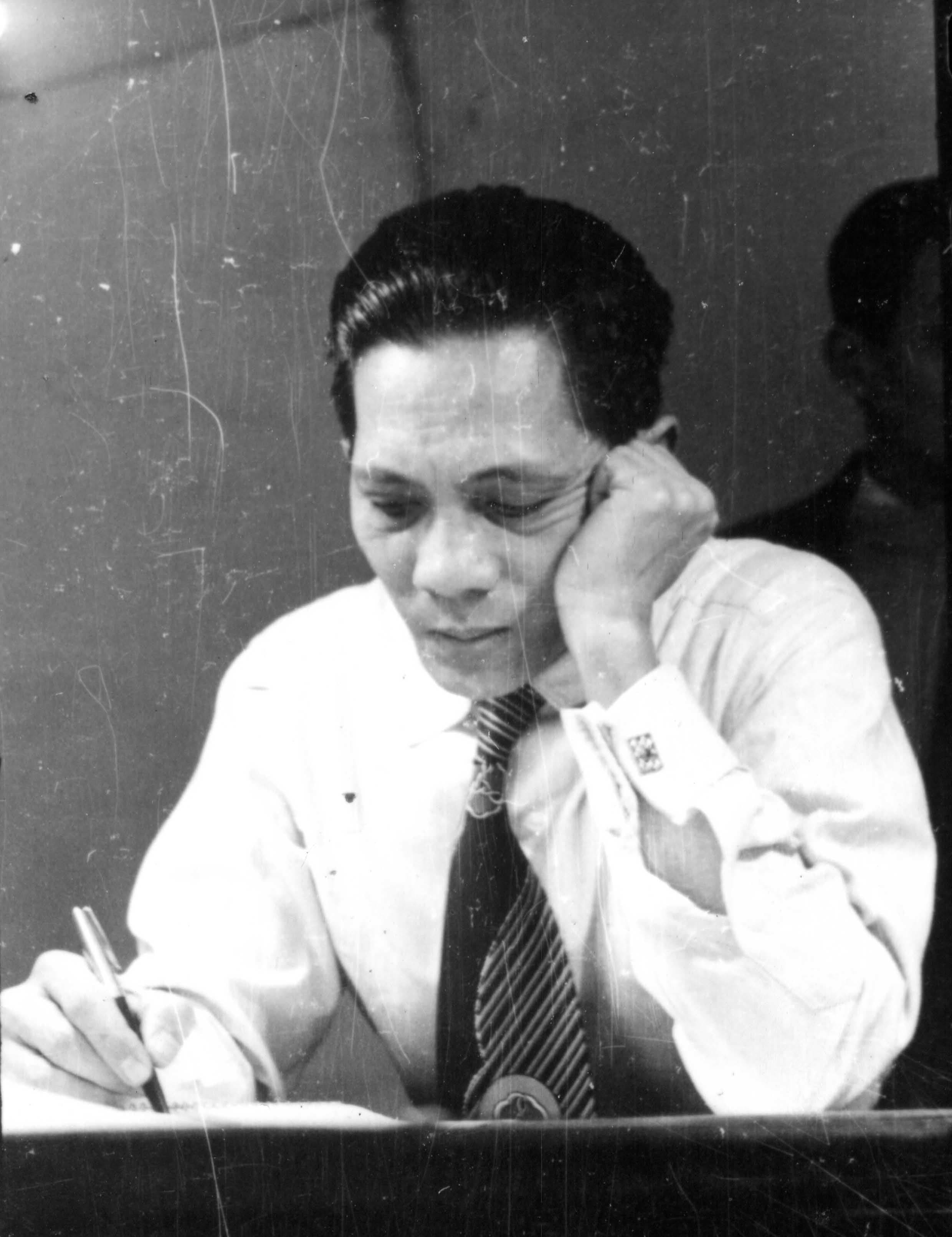 Man in a white shirt and striped tie writing at a desk, hand on his head.