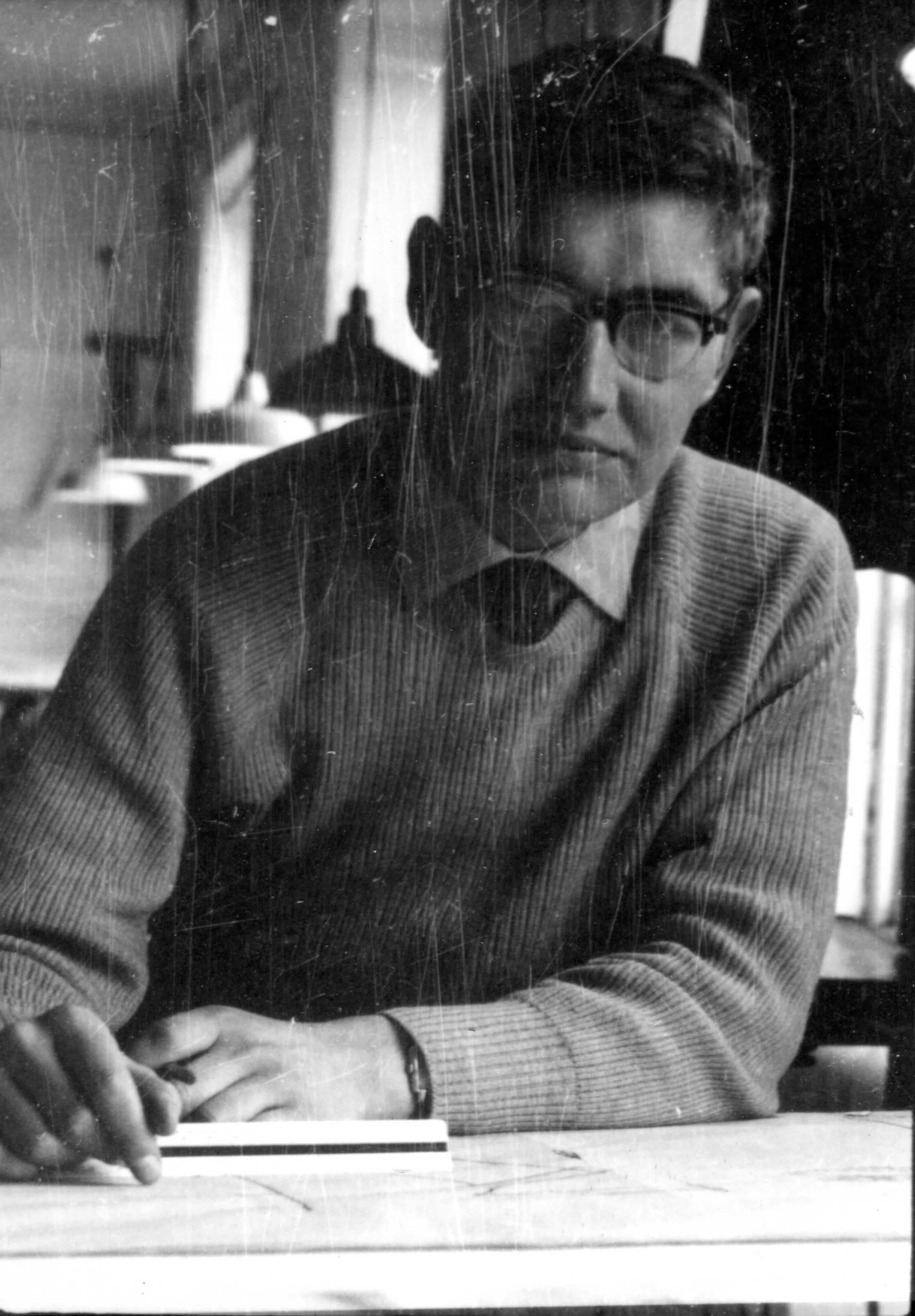 Man in glasses, sweater, and shirt, sitting at a desk looking at the viewer.
