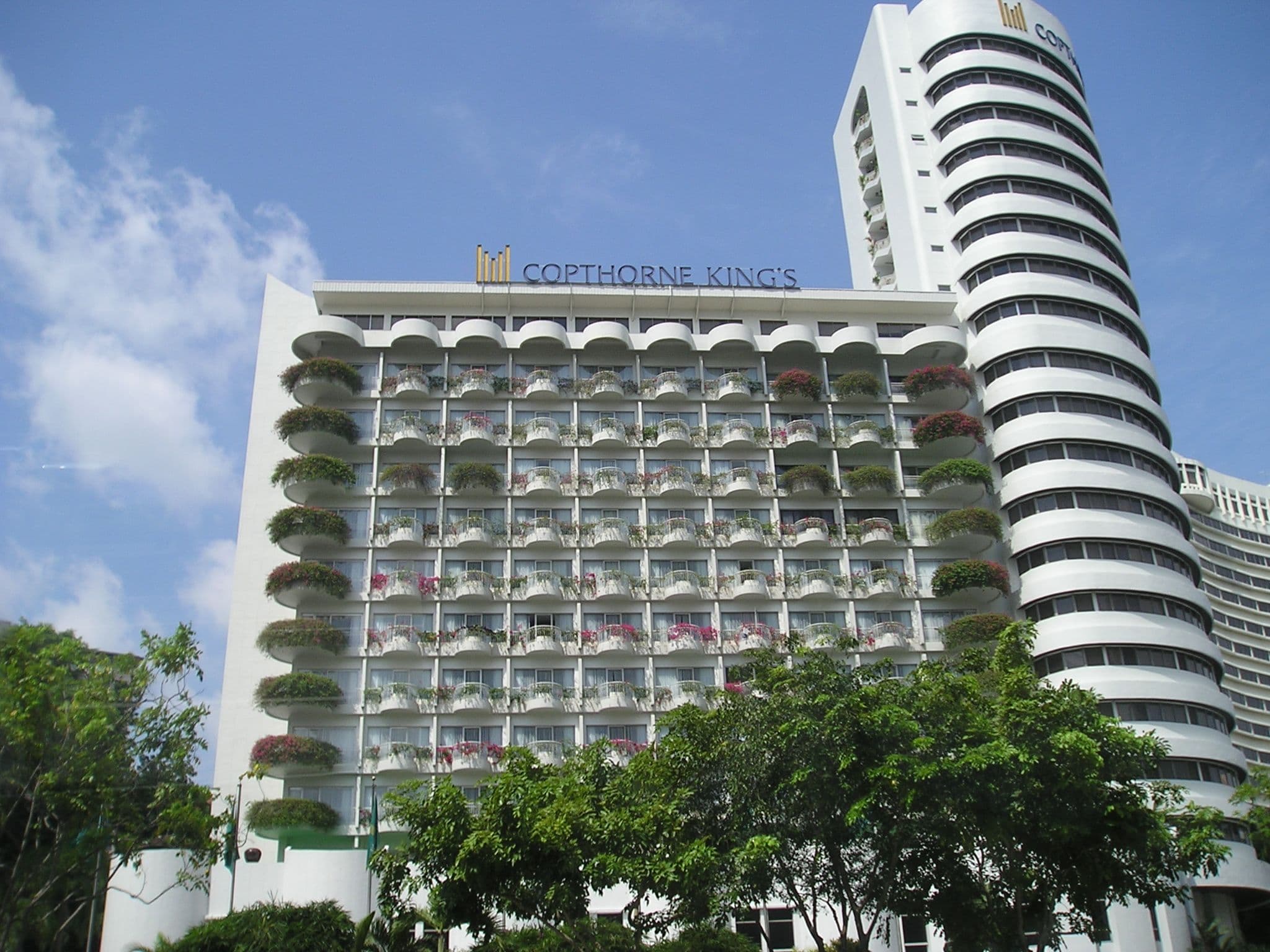 Tall white building with many balconies and a curved tower, under a blue sky with clouds.