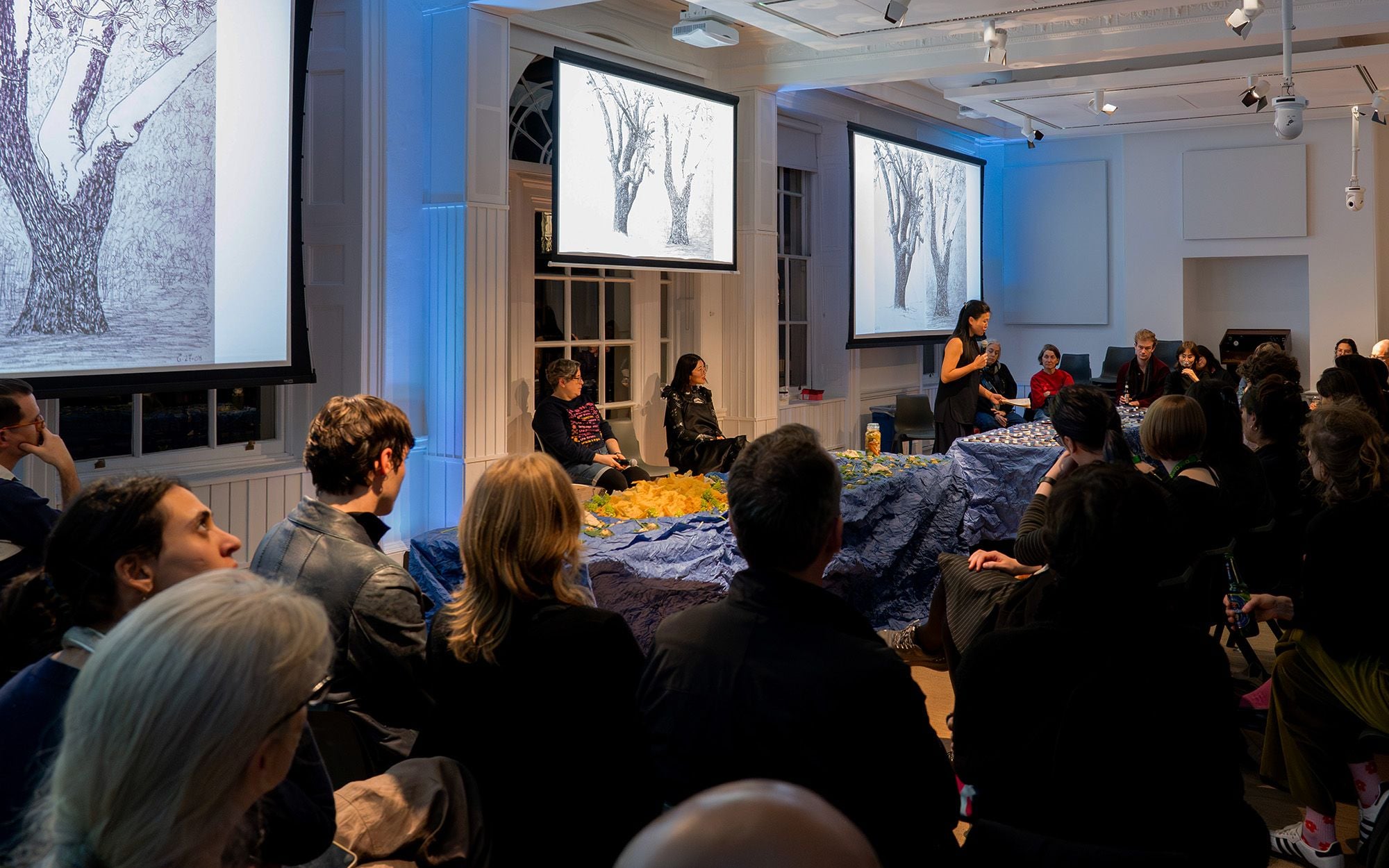 The Dream Maps event in the Lecture Hall with blue lights and people gathered around two tables with an installion of food as a landscape