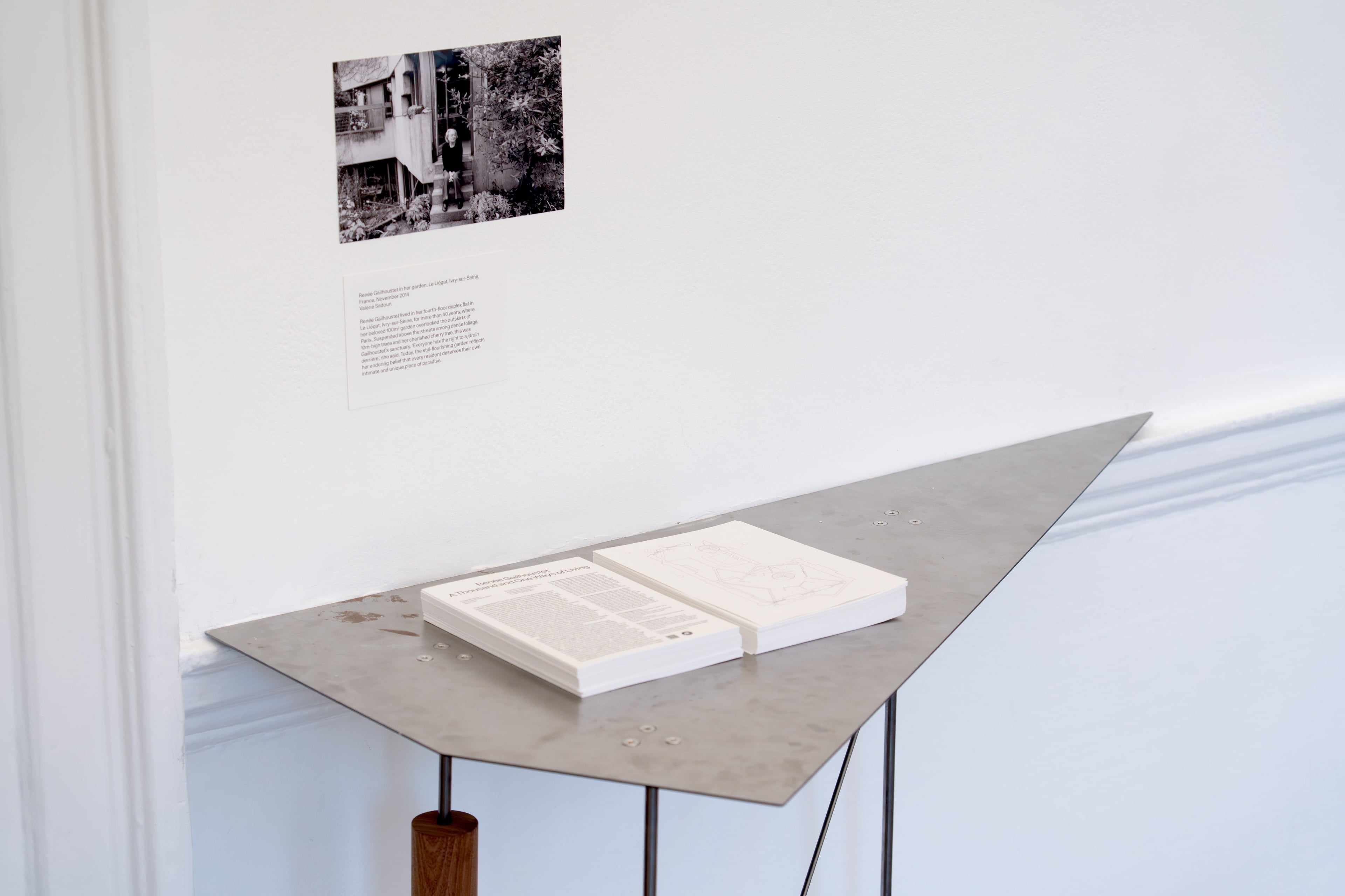 White paper exhibition guides are stacked on a triangular metal table below a white wall with a photo print on it.