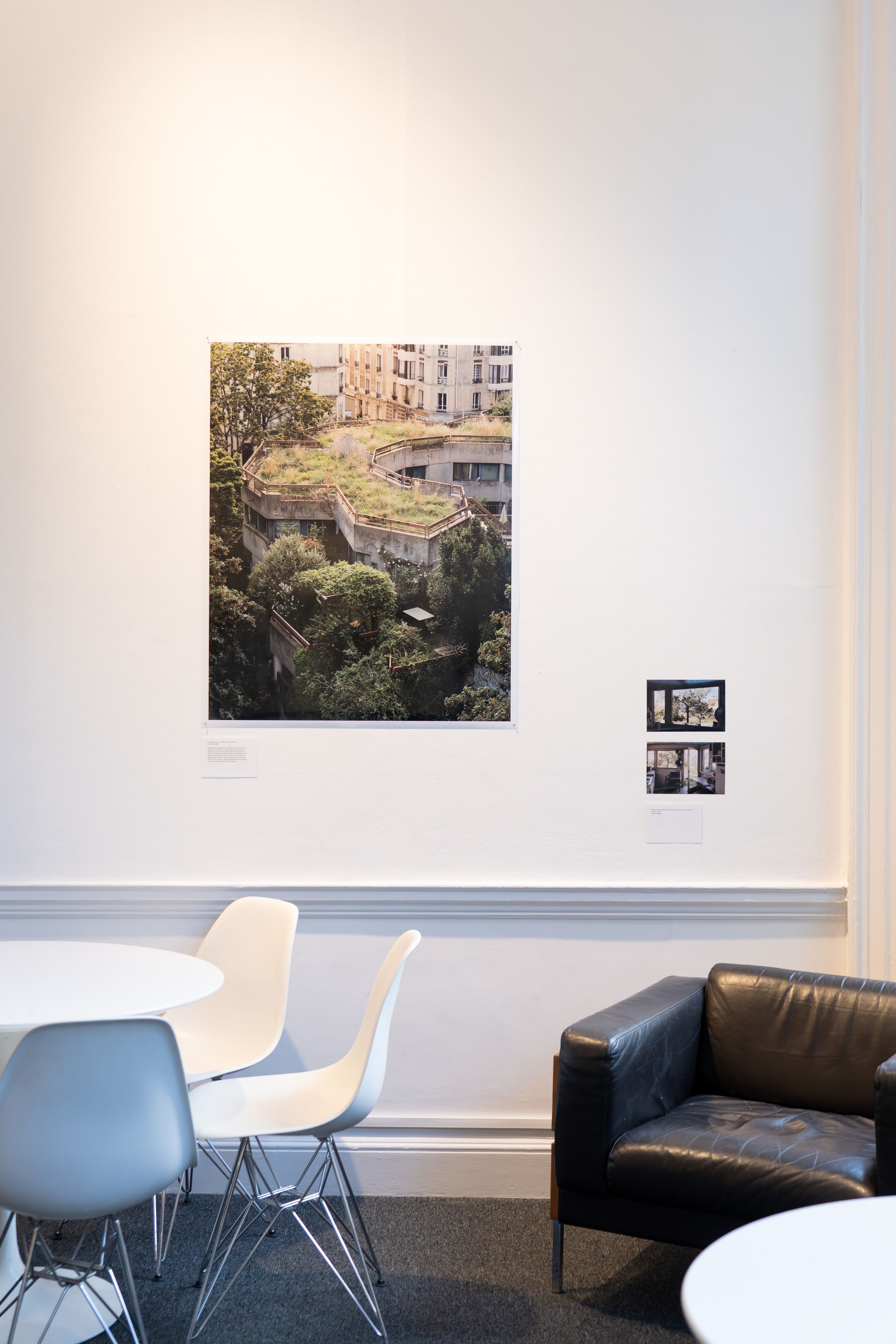 A photo of a building with roof garden is mounted on a white wall, with chairs and table in the foreground and a leather sofa.