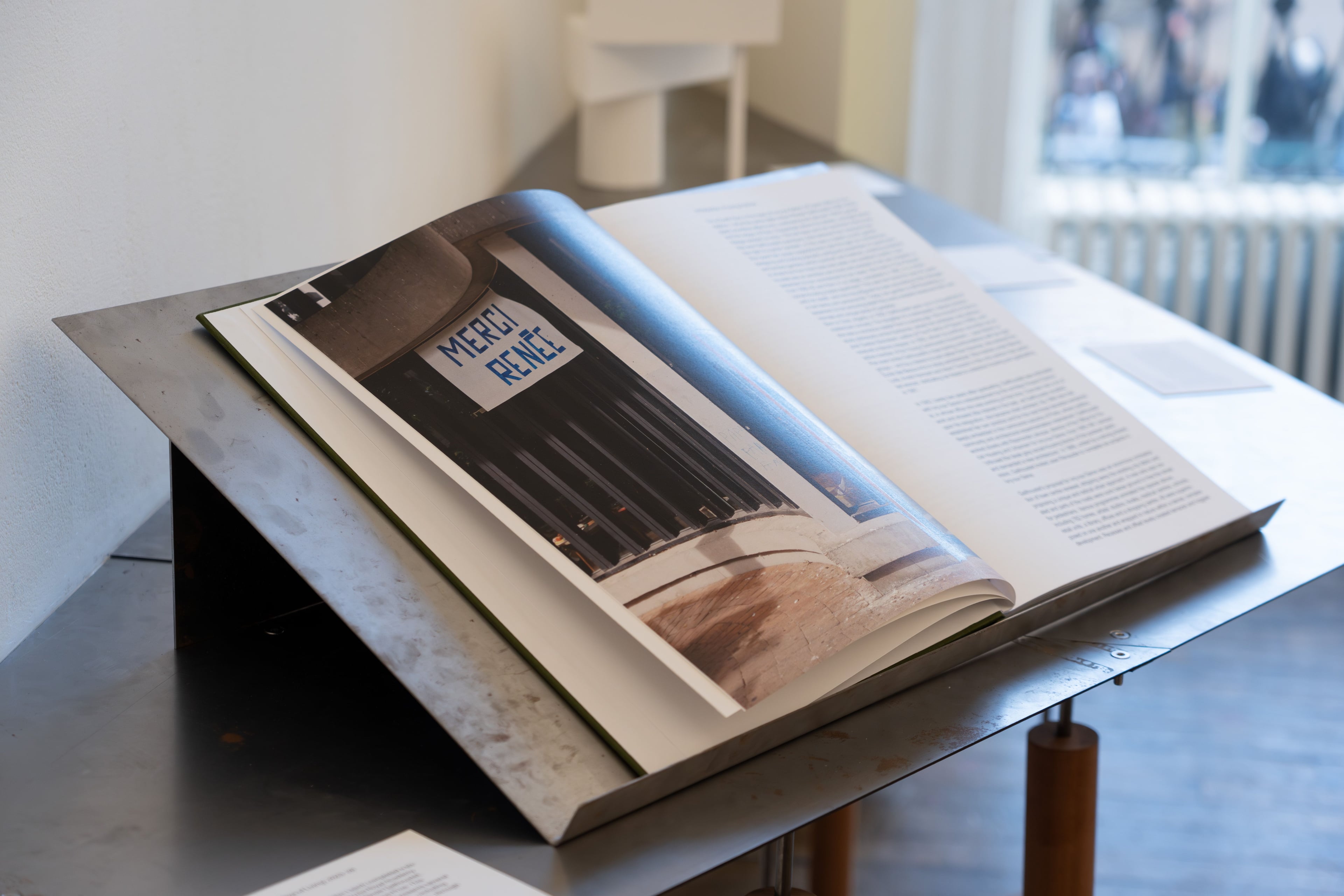 A large book of photos and text sits open on a metal book stand in the gallery.