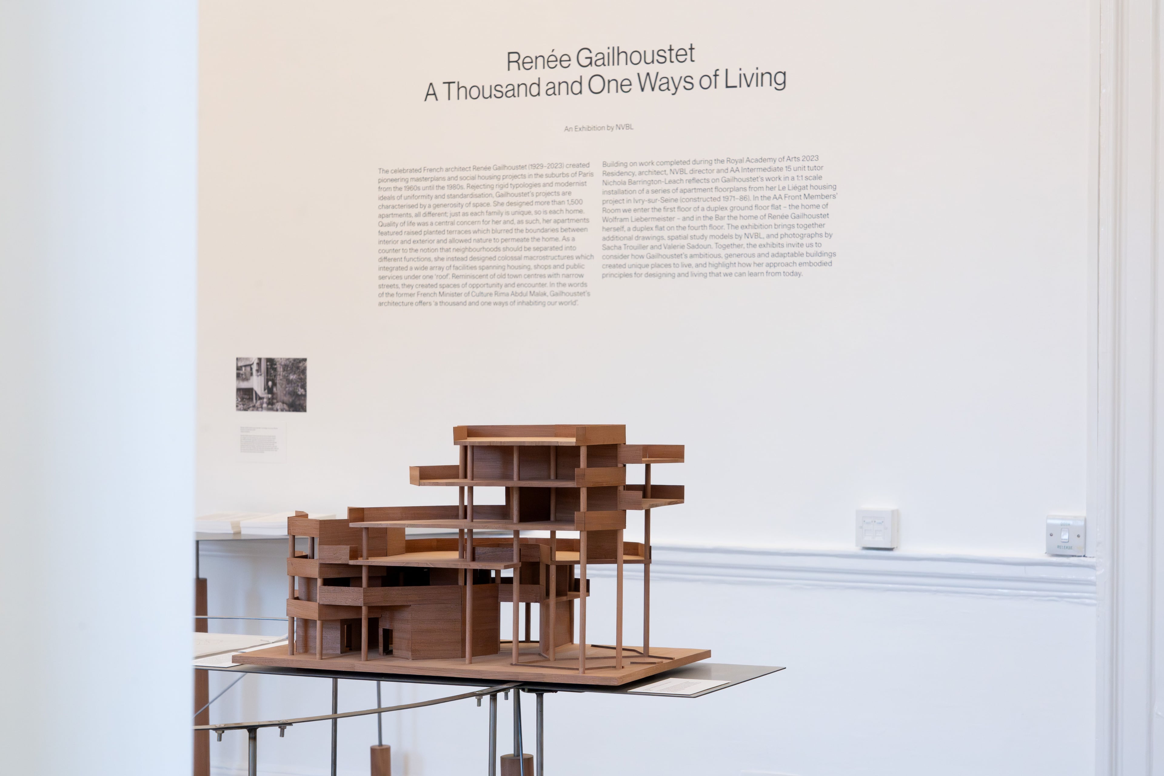 A wooden model sits on a metal table against a white wall with exhibition text.