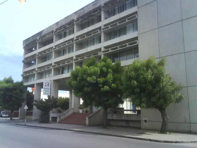 Modern concrete building with shaded balconies and green trees along a street.