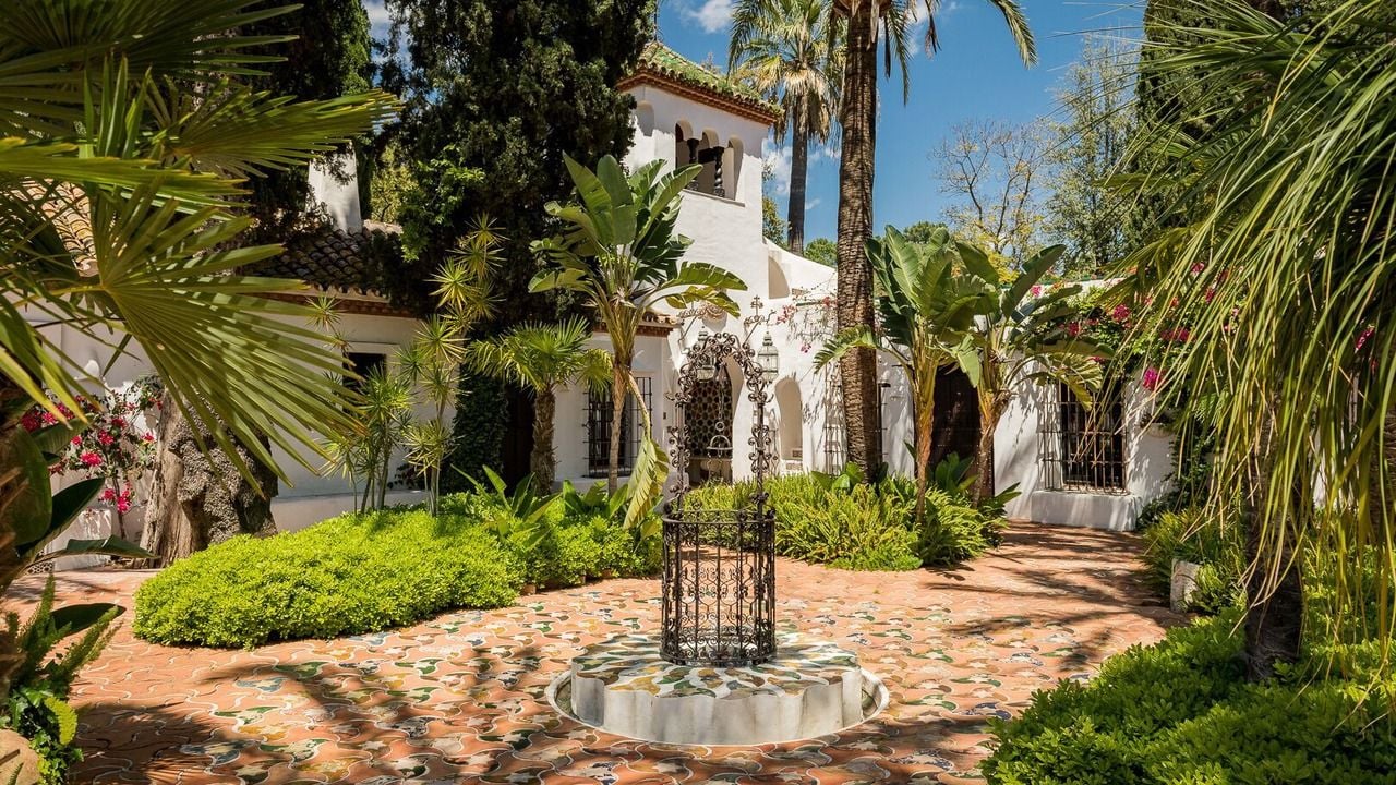 White stucco villa, lush palm tree garden, brick courtyard, and decorative well.