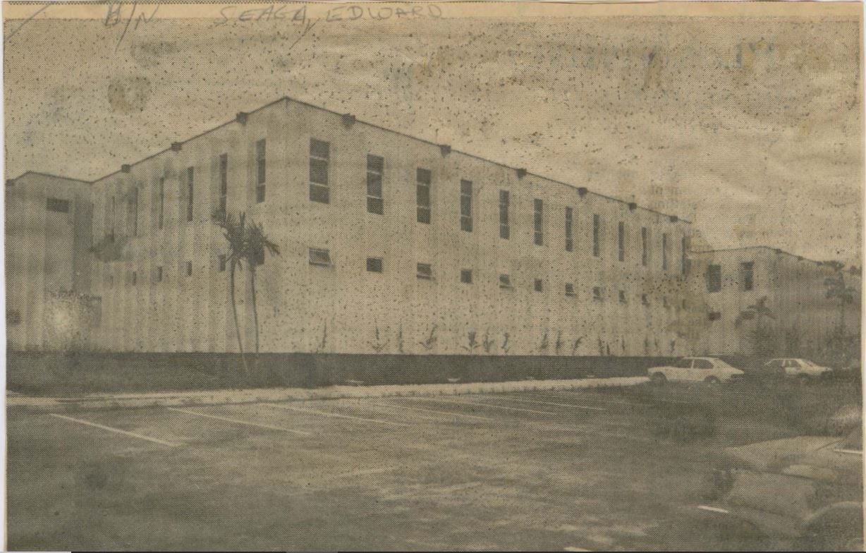 Exterior view of a long, two-story, light-colored building with many windows and a fenced perimeter.