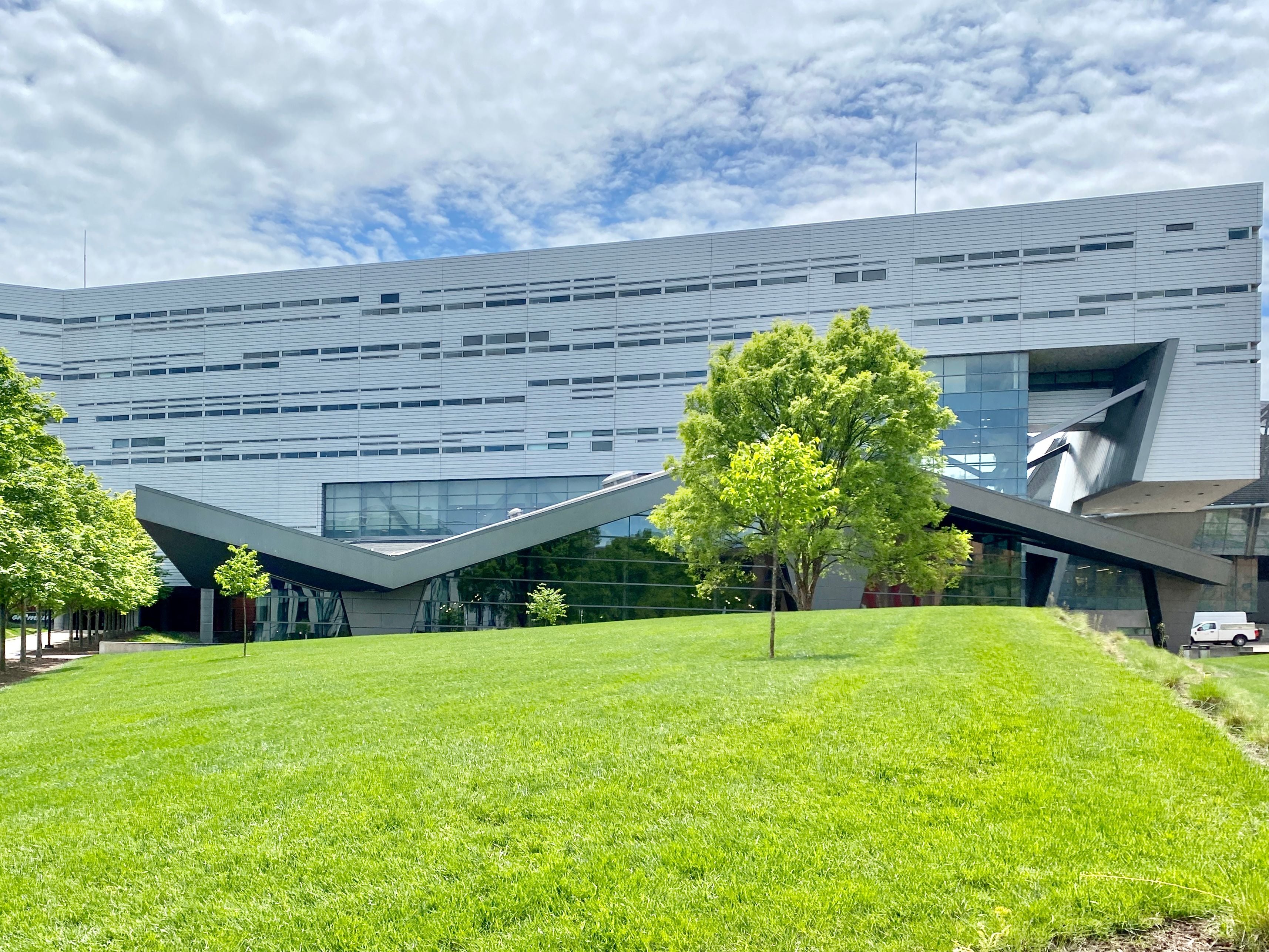 Modern geometric building with glass and metal facade surrounded by green lawn and trees.
