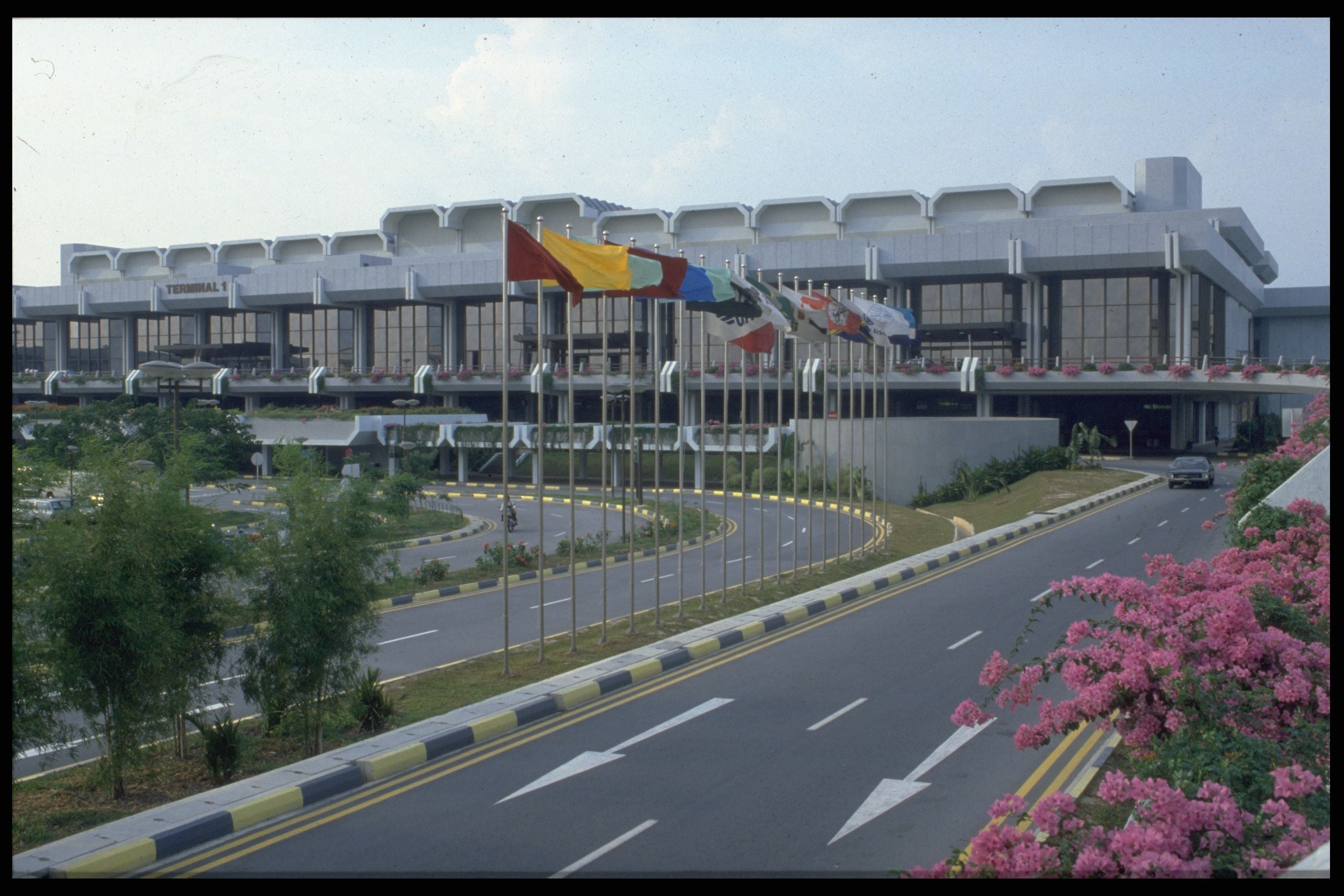 Modern airport terminal building with flags, a curved road, and landscaping under a clear sky.