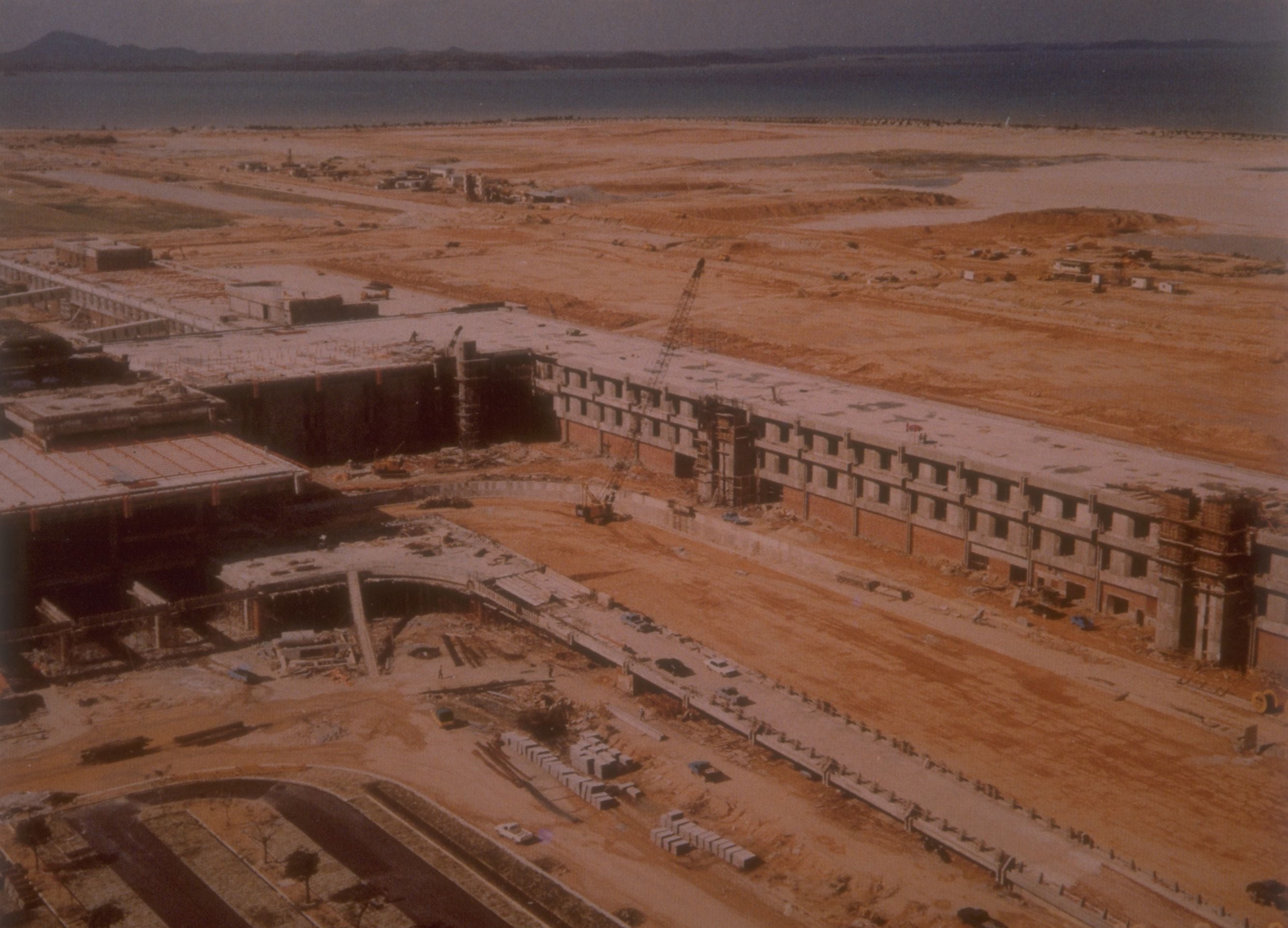 Aerial view of a large building under construction on a sandy coast beside blue water.