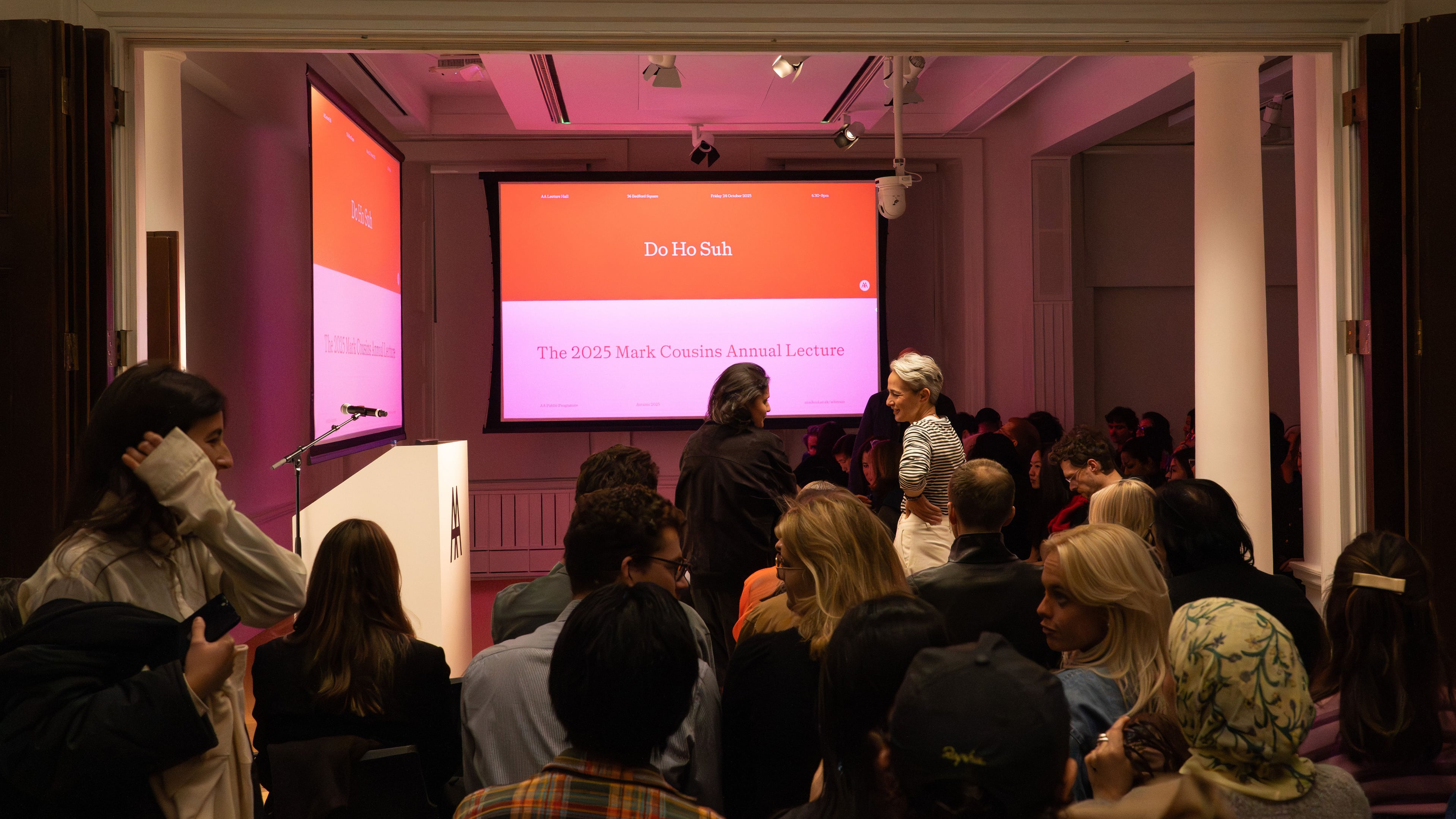 A dimly lit room with a presentation screen displaying text and a crowd watching.