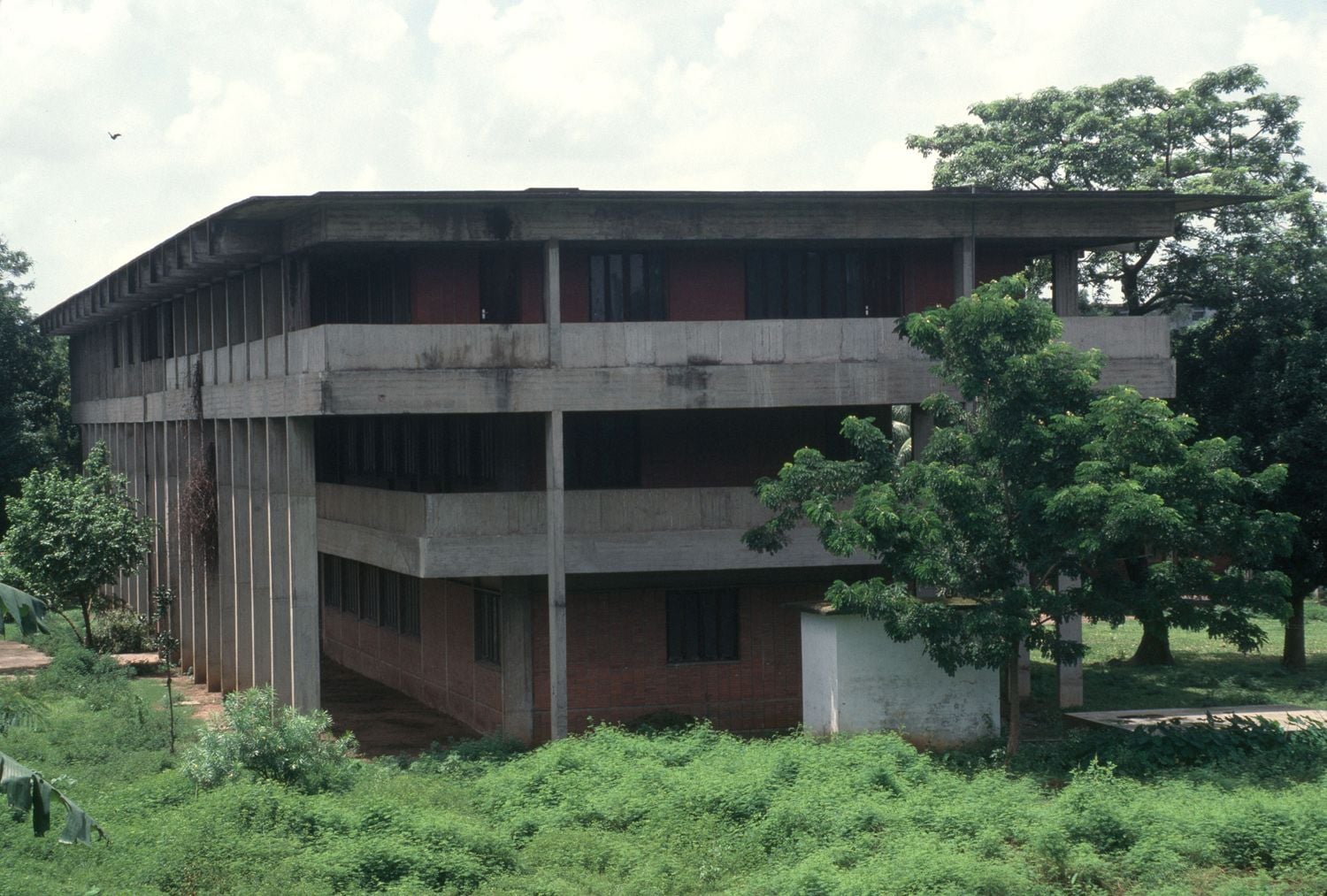 Brutalist concrete building with multiple levels and balconies surrounded by lush green foliage.