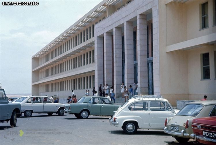 A row of vintage cars parked in front of a modern, multi-story building with a colonnaded entrance.