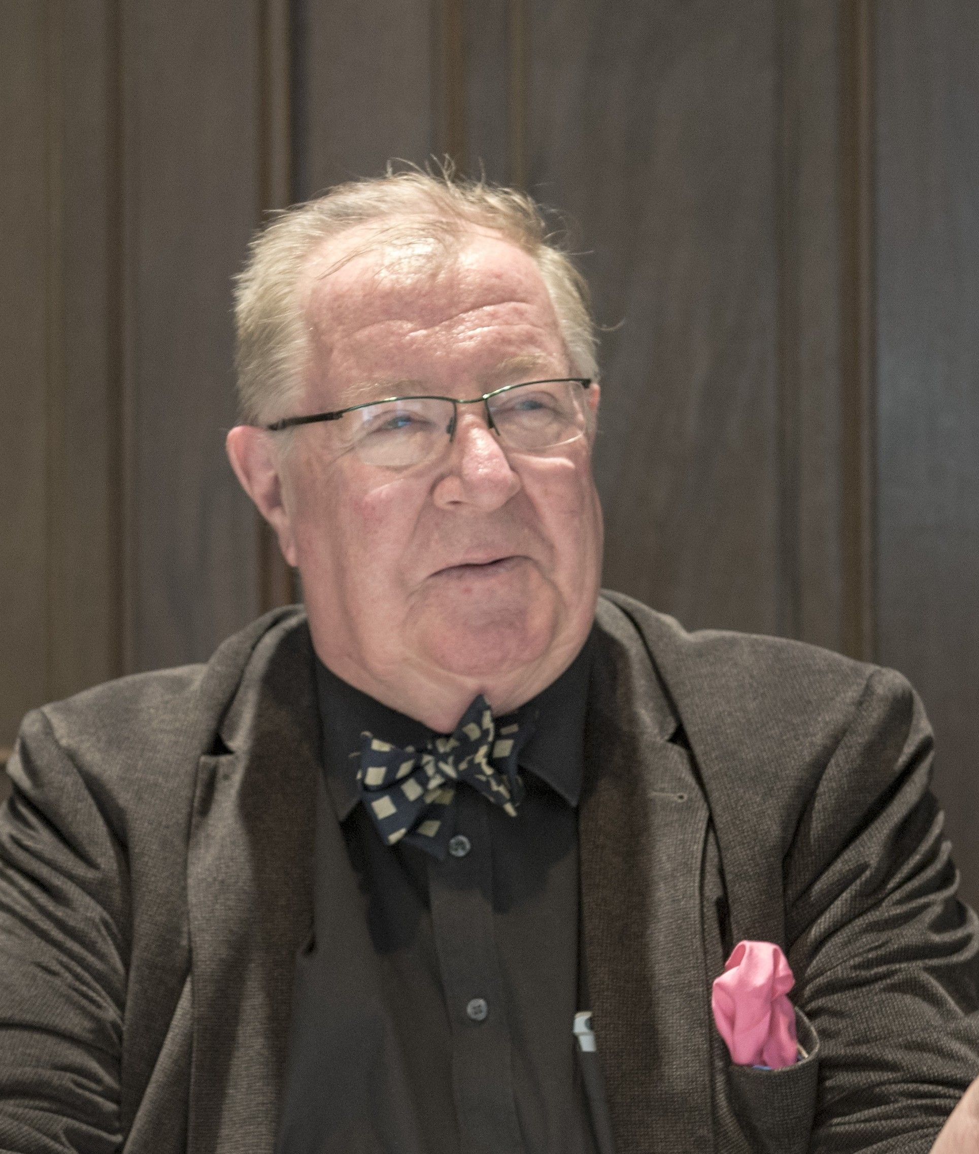 Older man with glasses, bow tie, and pink pocket square sitting at a table.