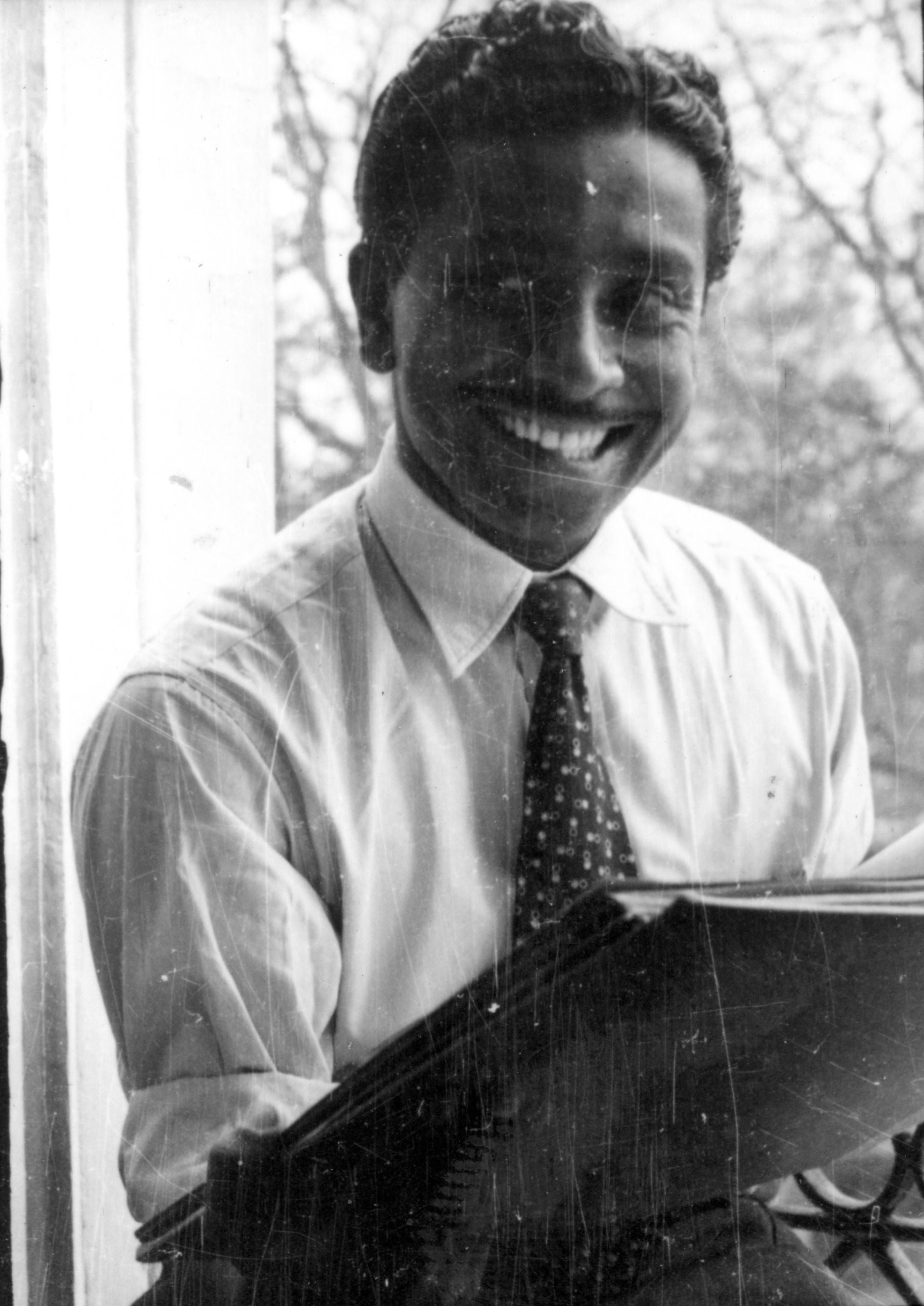 A smiling Black man with a mustache wears a collared shirt and tie while sitting at a desk.