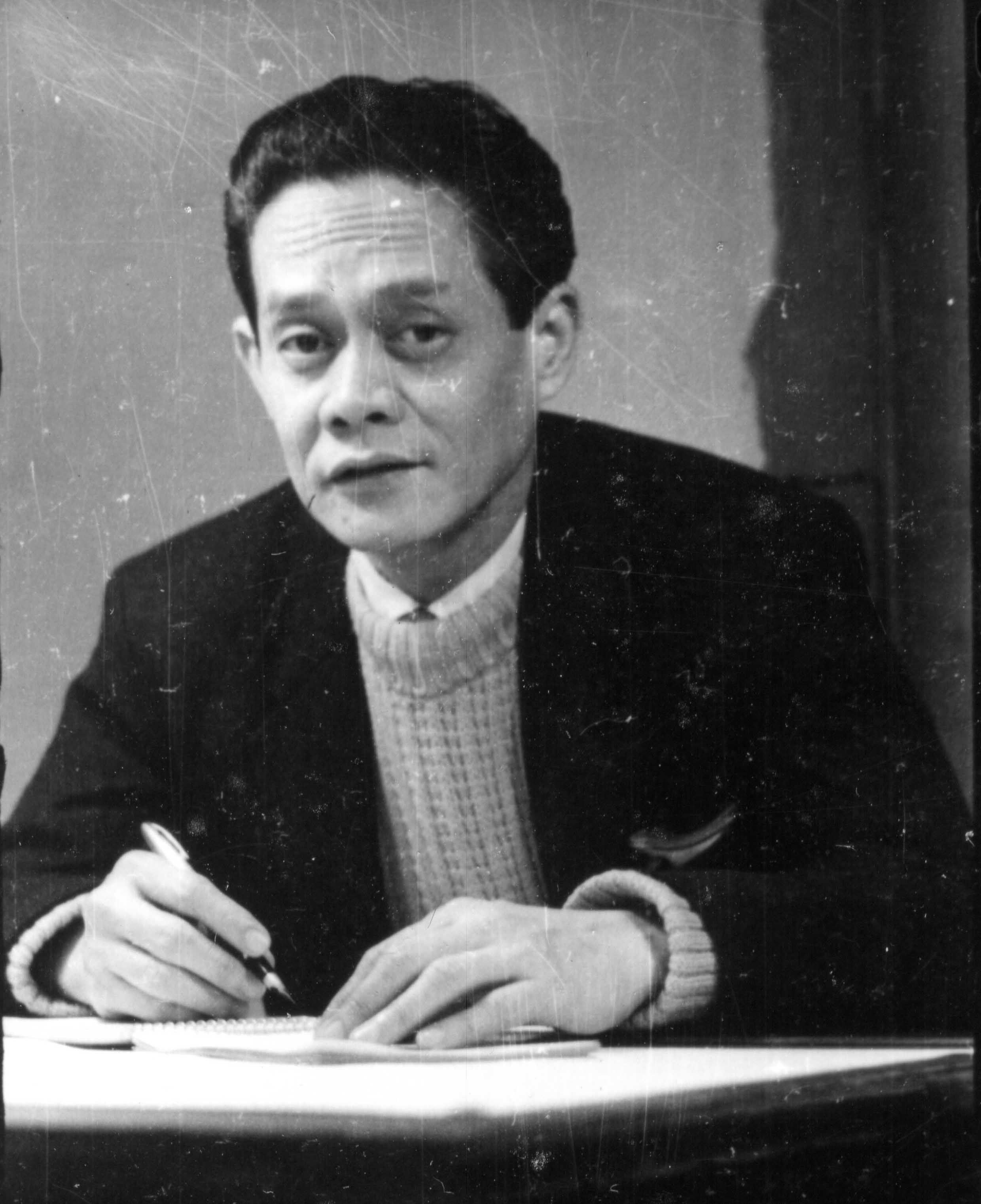 Man in a dark jacket and sweater writing at a desk, looking thoughtfully to the side.