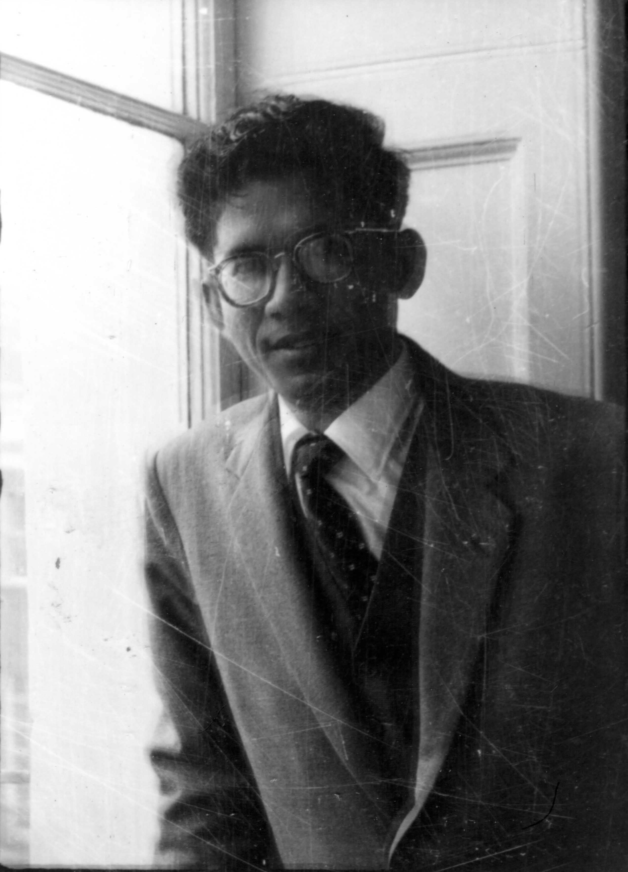 Man in glasses, suit, and tie sitting by a window, looking thoughtfully away.