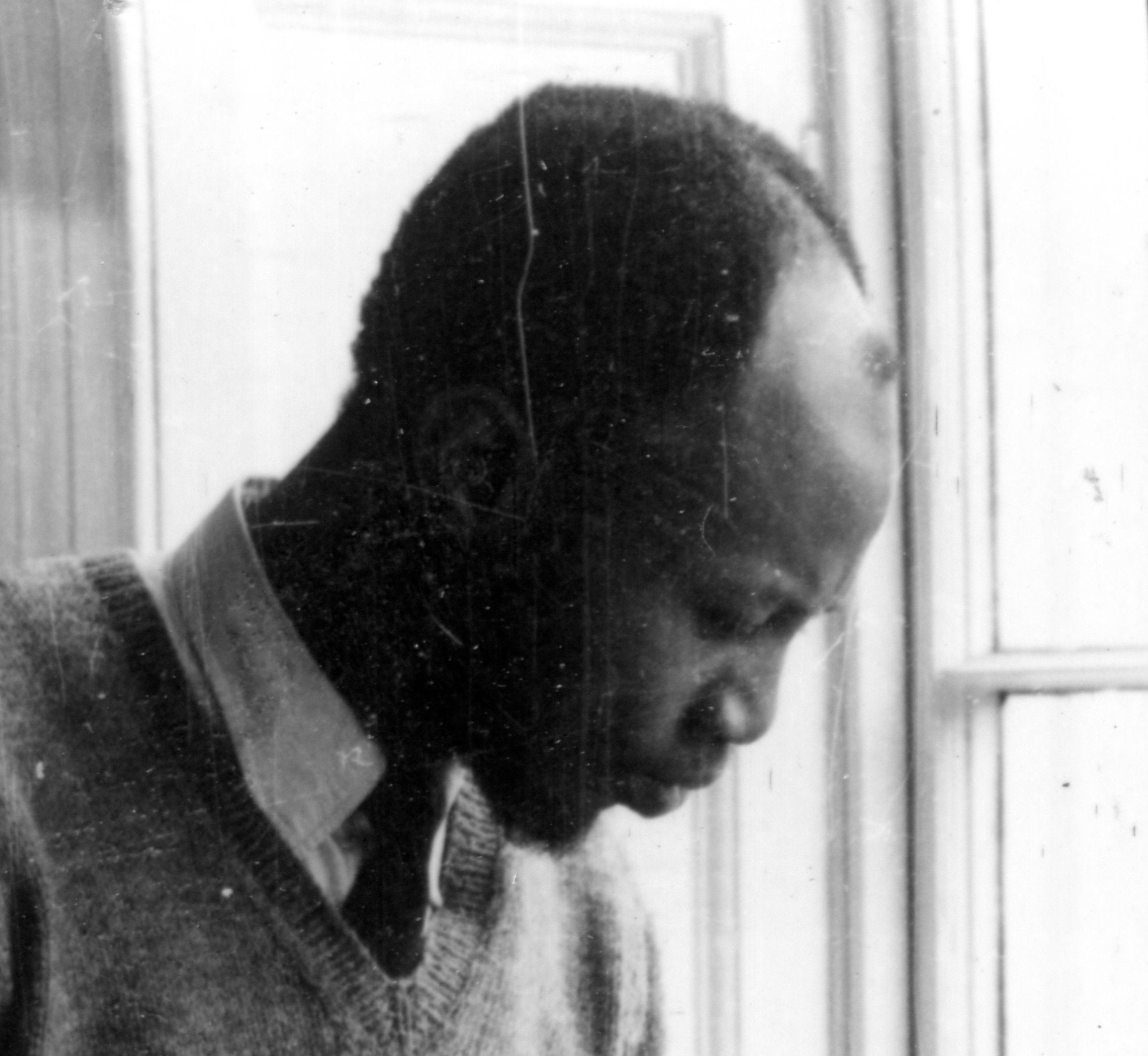 A man in a sweater vest and collared shirt looks down thoughtfully near a window.