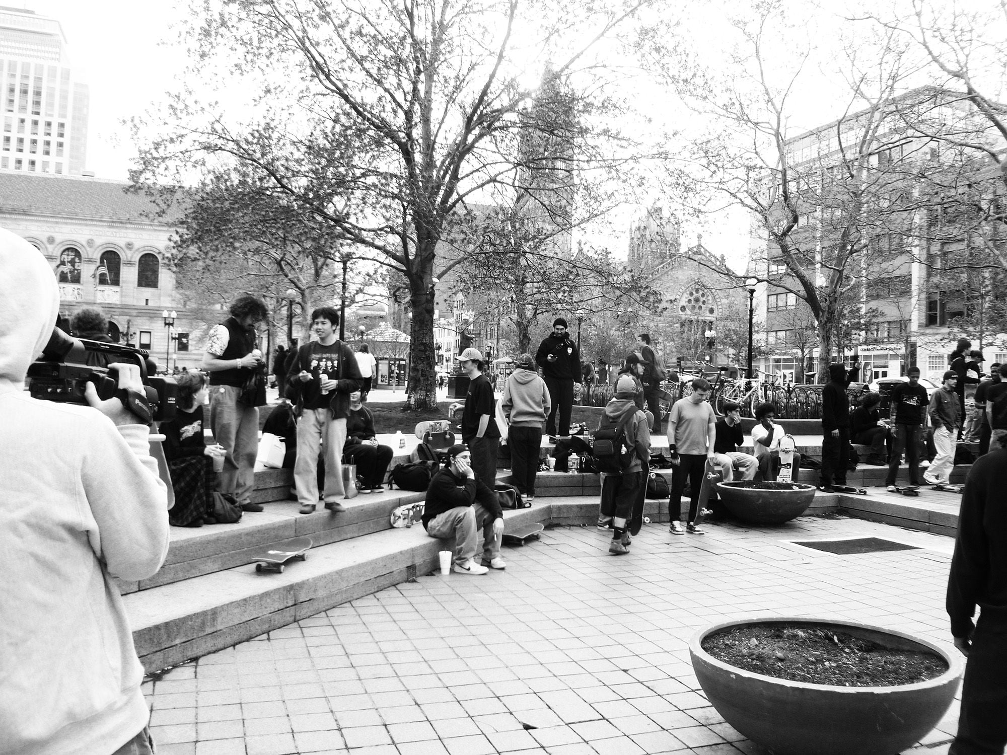 A group of people stand, sit and skateboard in a public square with steps and trees