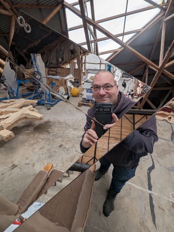 Man in glasses holds up a reflective surface in a workshop.