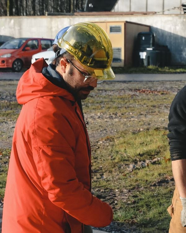 A man wearing a yellow hard hat and a bright orange jacket stands in a working yard.