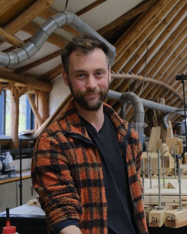 A white skinned man with brown hair and a short beard stands in a workshop surrounded by woodworking.