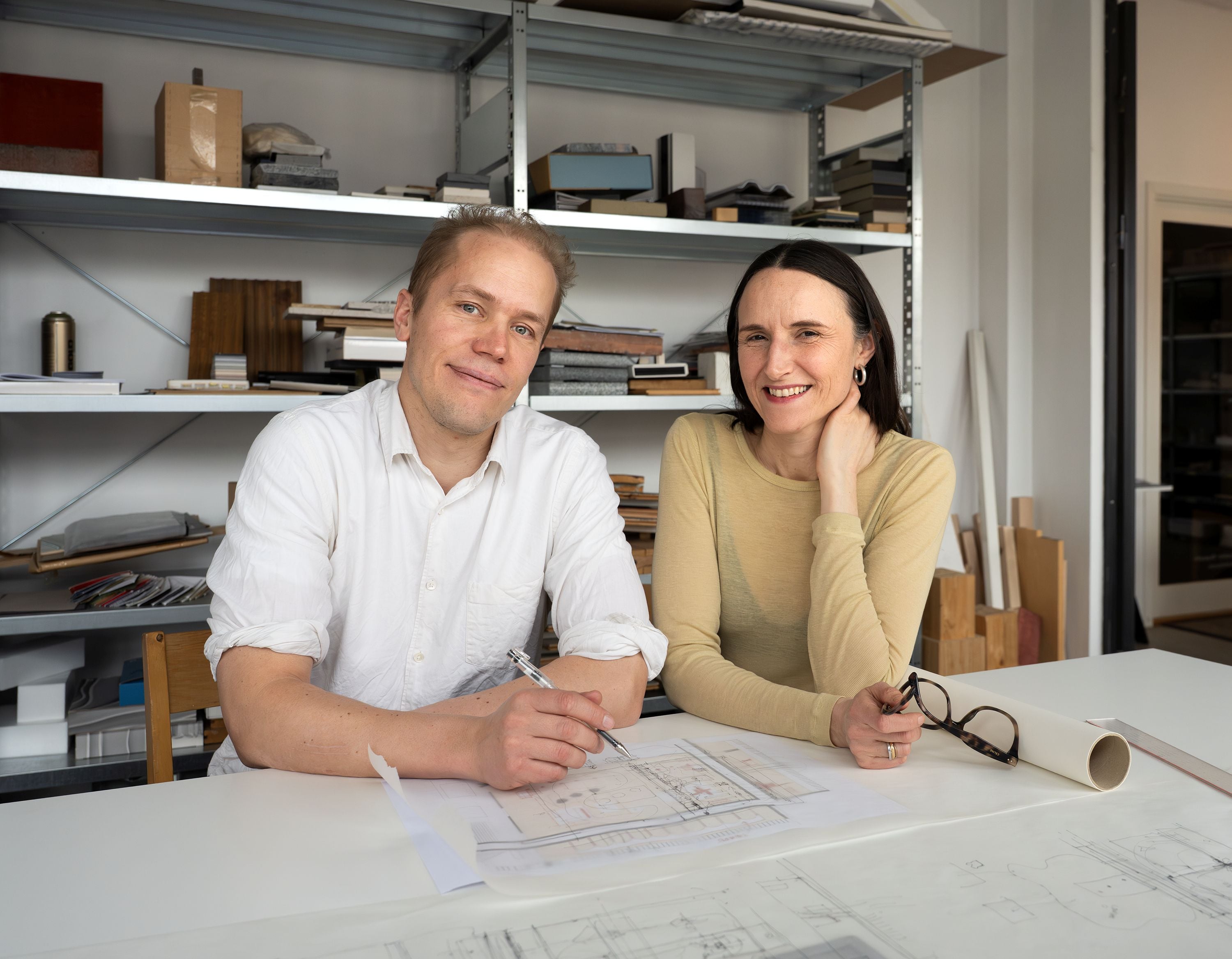 Photograph of two people smiling at the camera who are sat at a desk with architectural drawings in front of them