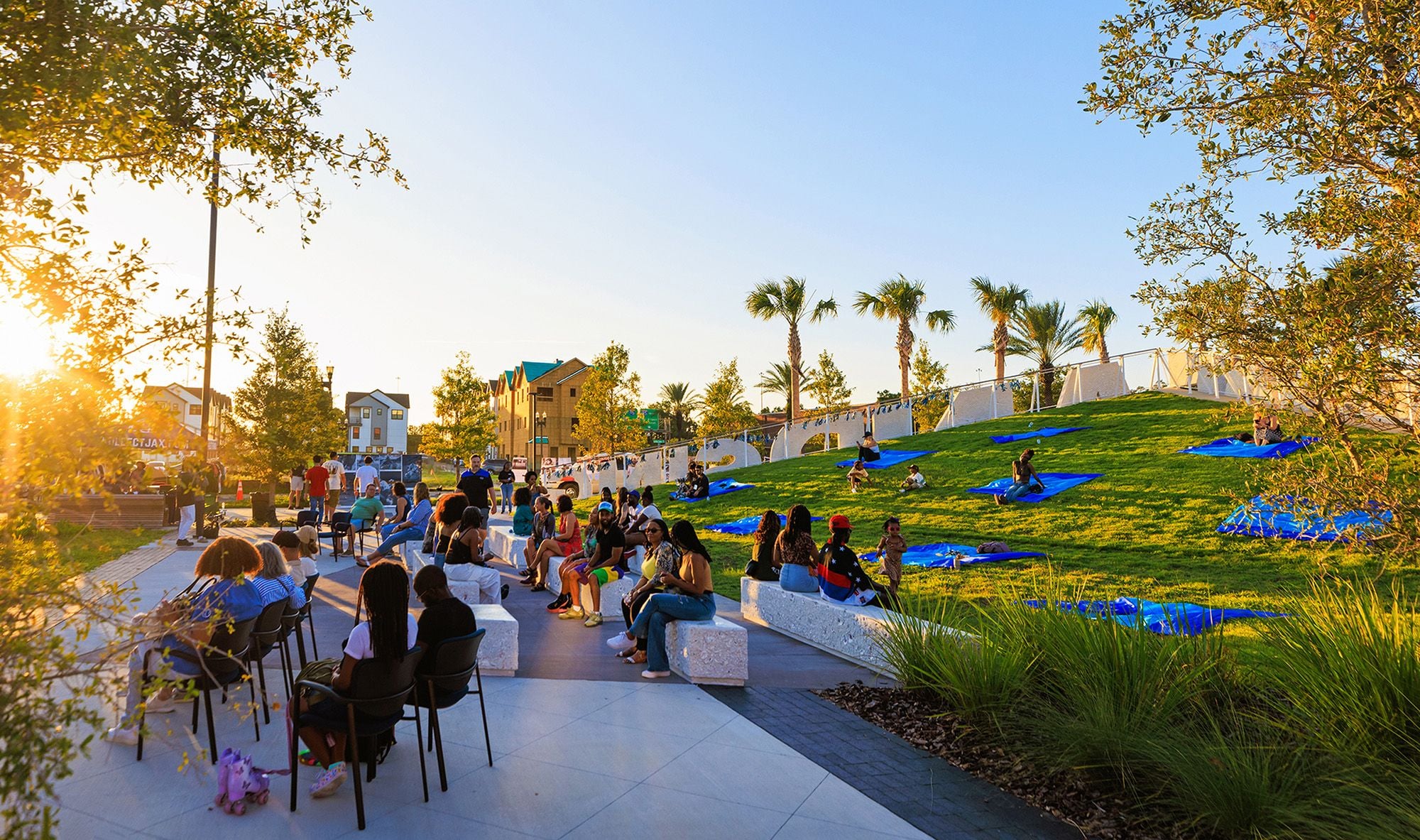 A public park with golden evening light shining on people sitting on benches and chair surrounded by grass and trees