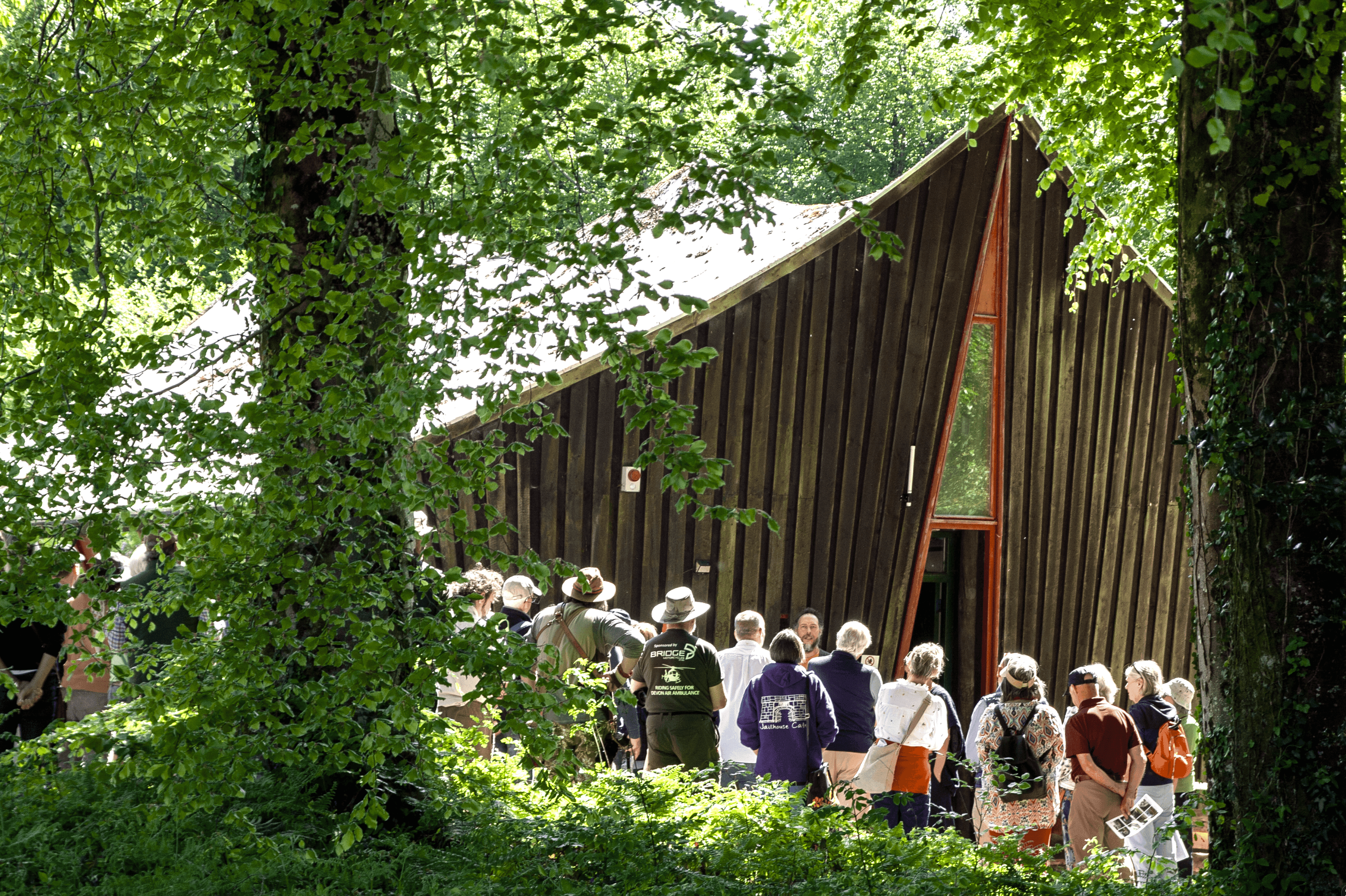 A crowd of people stand in front of a timber-clad building in a green forest.