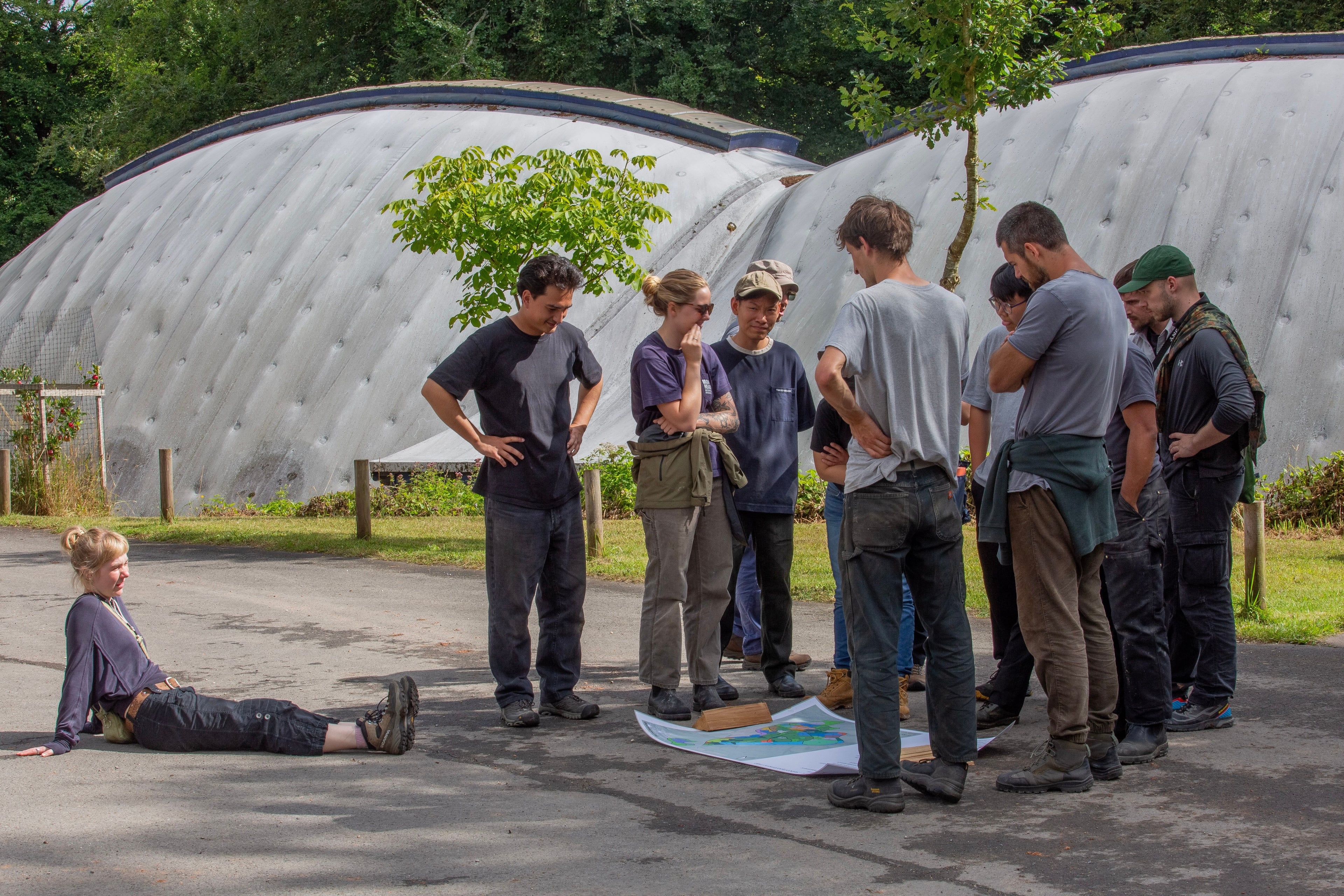 A group of people gather around a map on the ground in front of the Workshop at Hooke Park, while one person sits separately.