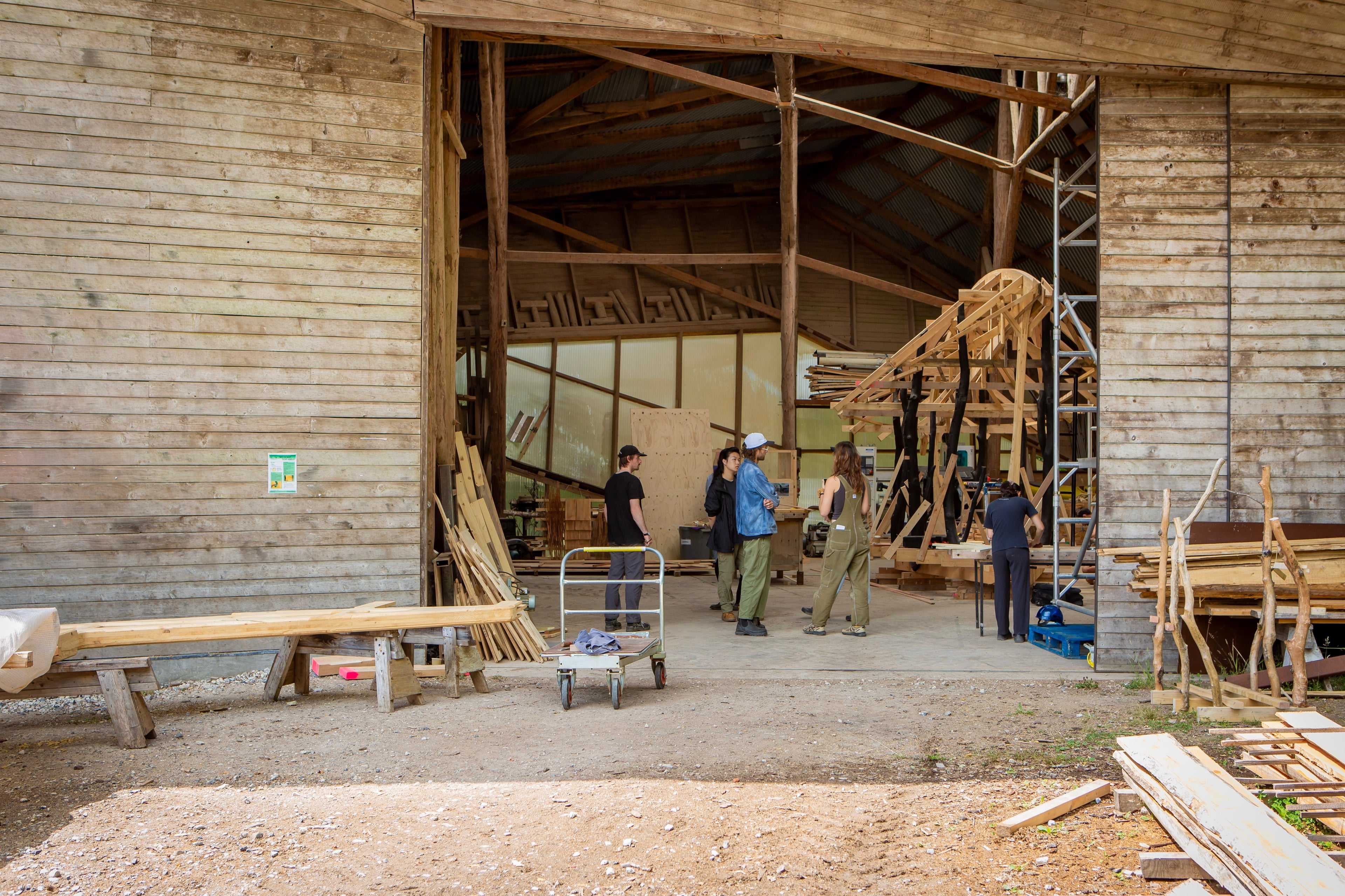 A group of people stand in the entrance to a huge wooden workshop, in front of a half-built timber structure.