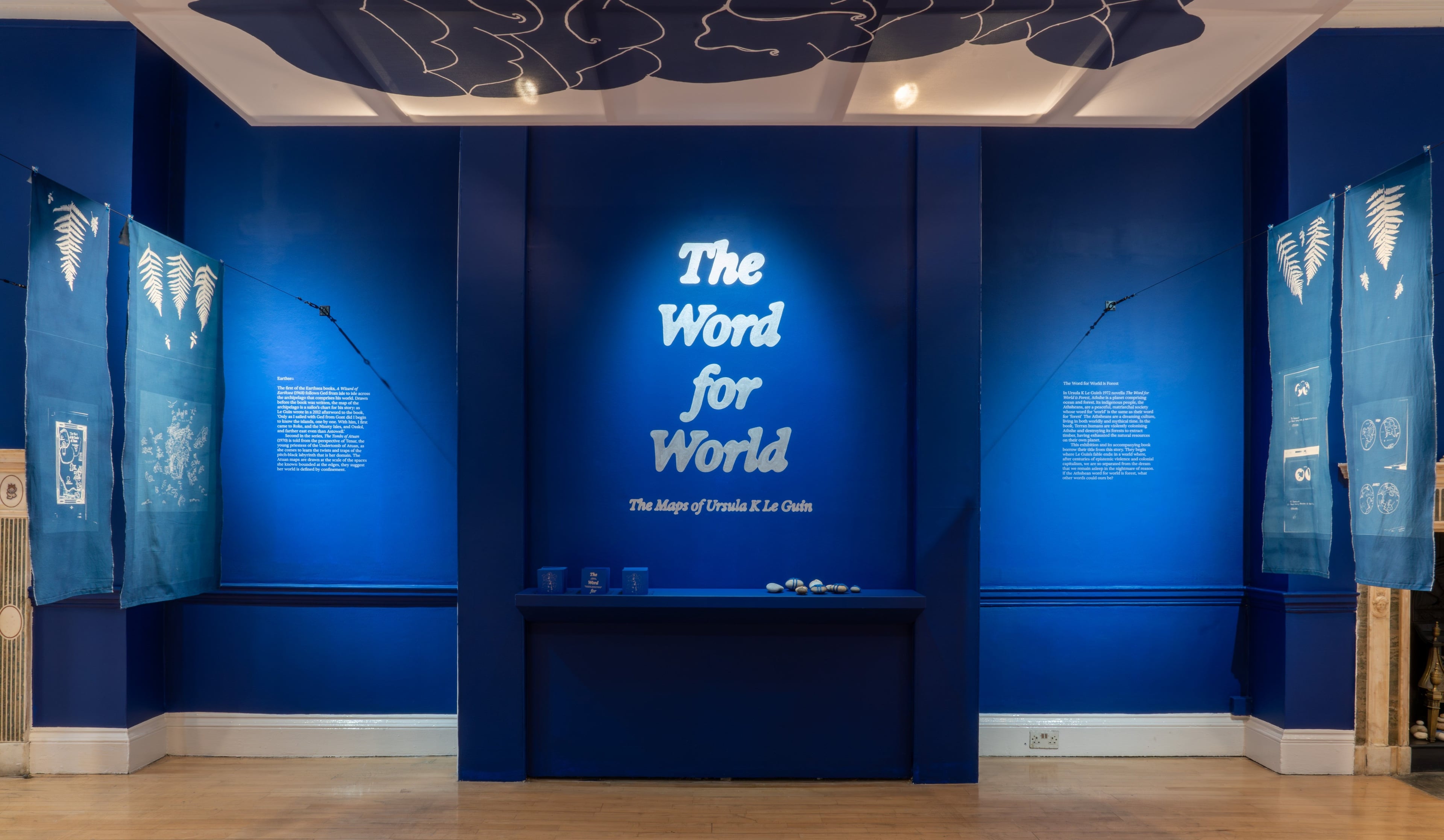 The Word for World exhibition with deep blue walls, a silver title and hanging blue cyanotype prints of maps