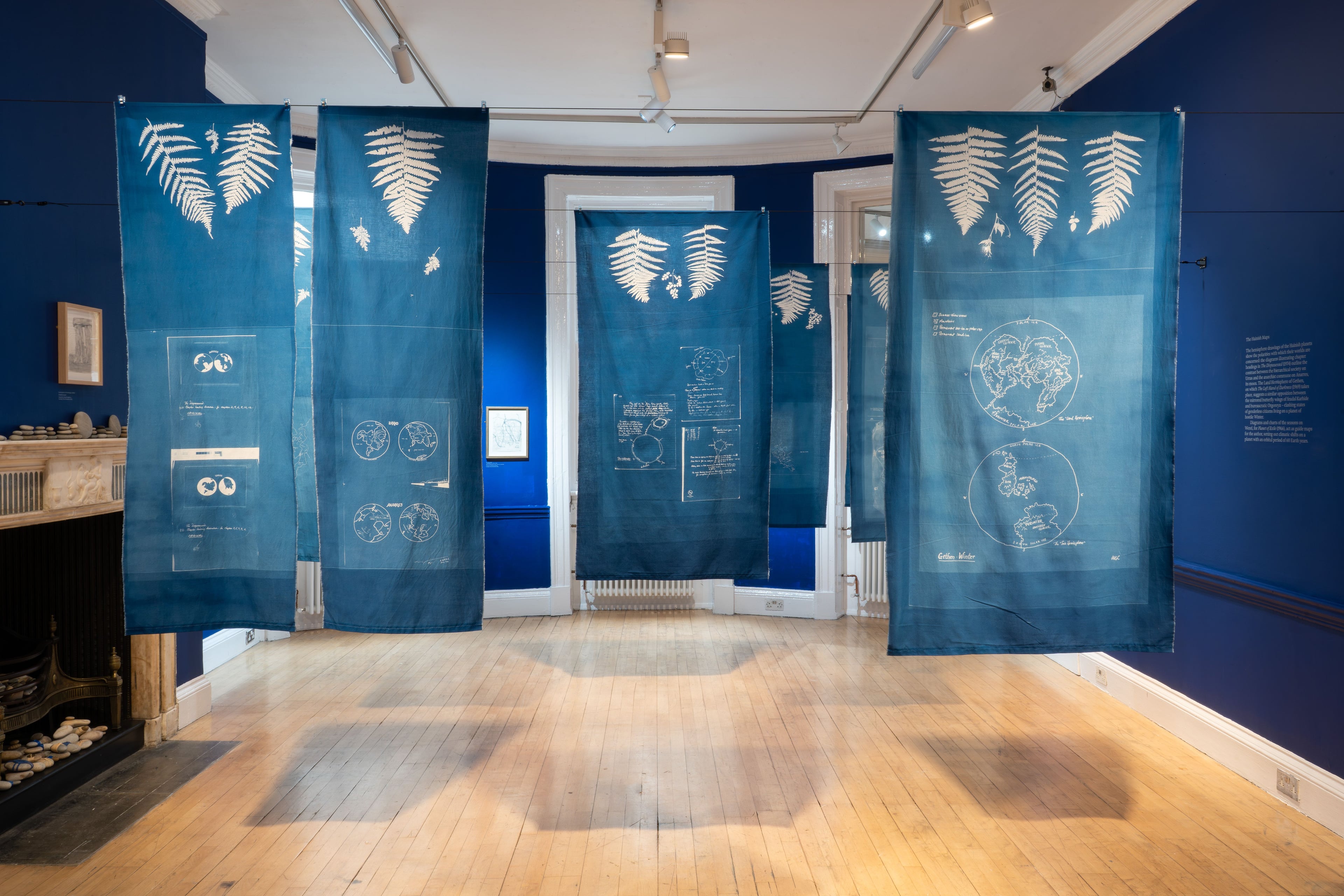 Installation shot of The Word for World exhibition showing the AA Gallery painted deep blue, with hanging fabric cyanotype prints of maps.